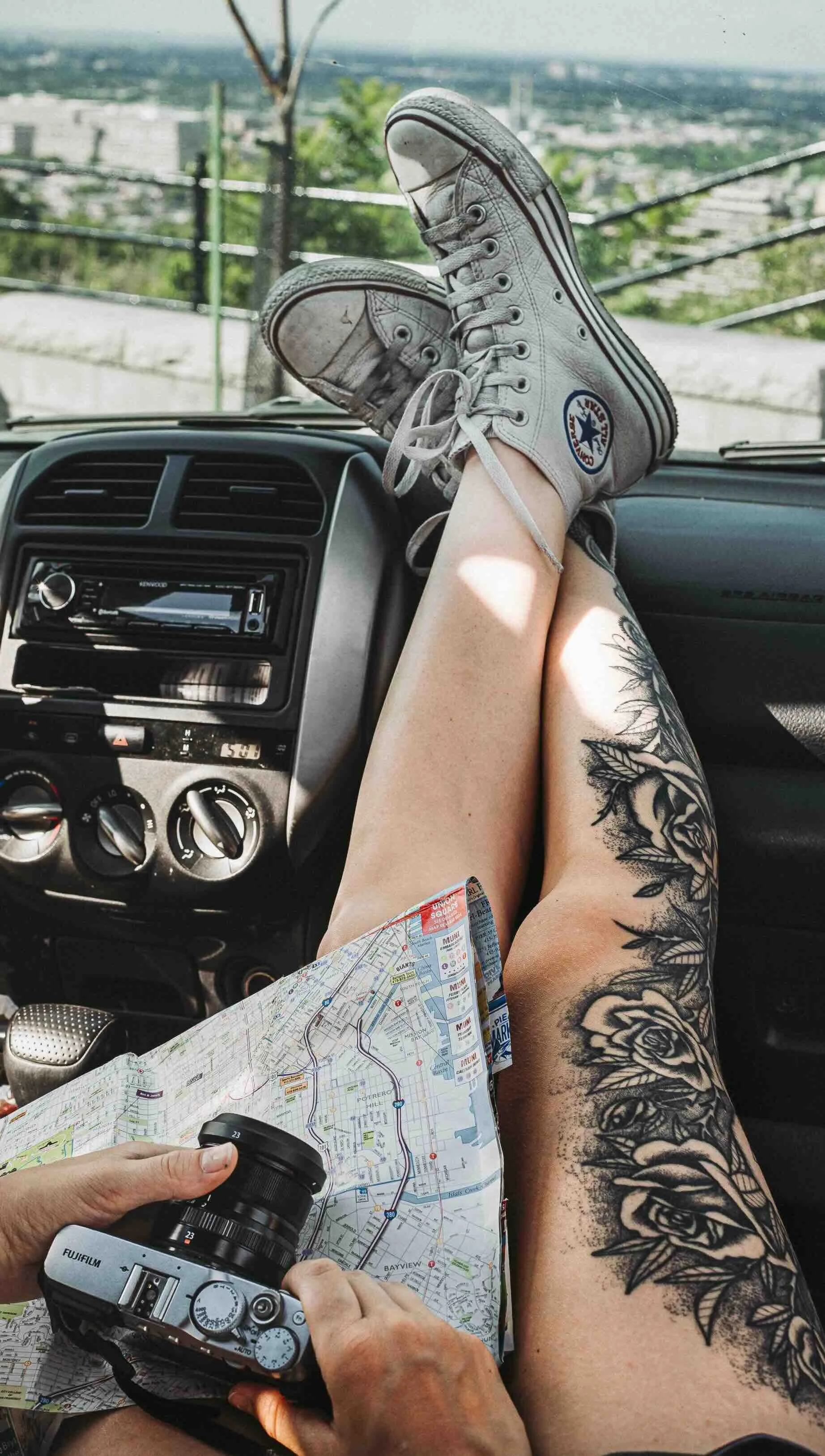 Personne avec une tatouage florale sur la jambe, portant des converses blanches, assise dans une voiture avec une carte et un appareil photo, vue extérieure de la voiture avec un paysage urbain en arrière-plan.