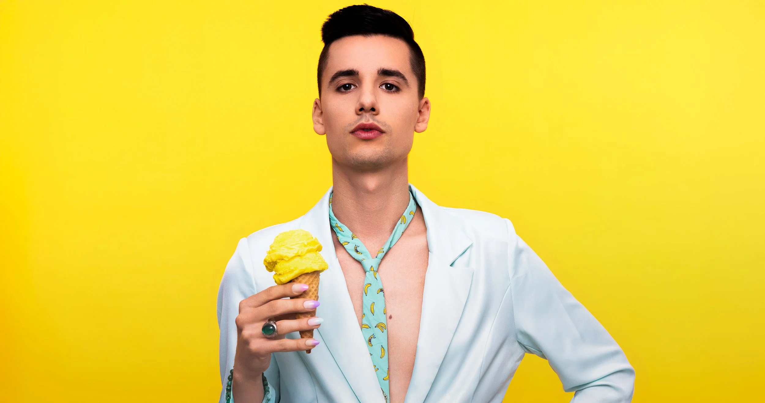 Jeune homme avec une chemise ouverte portant un foulard à motifs de bananes, tenant une glace à la pêche contre un fond jaune