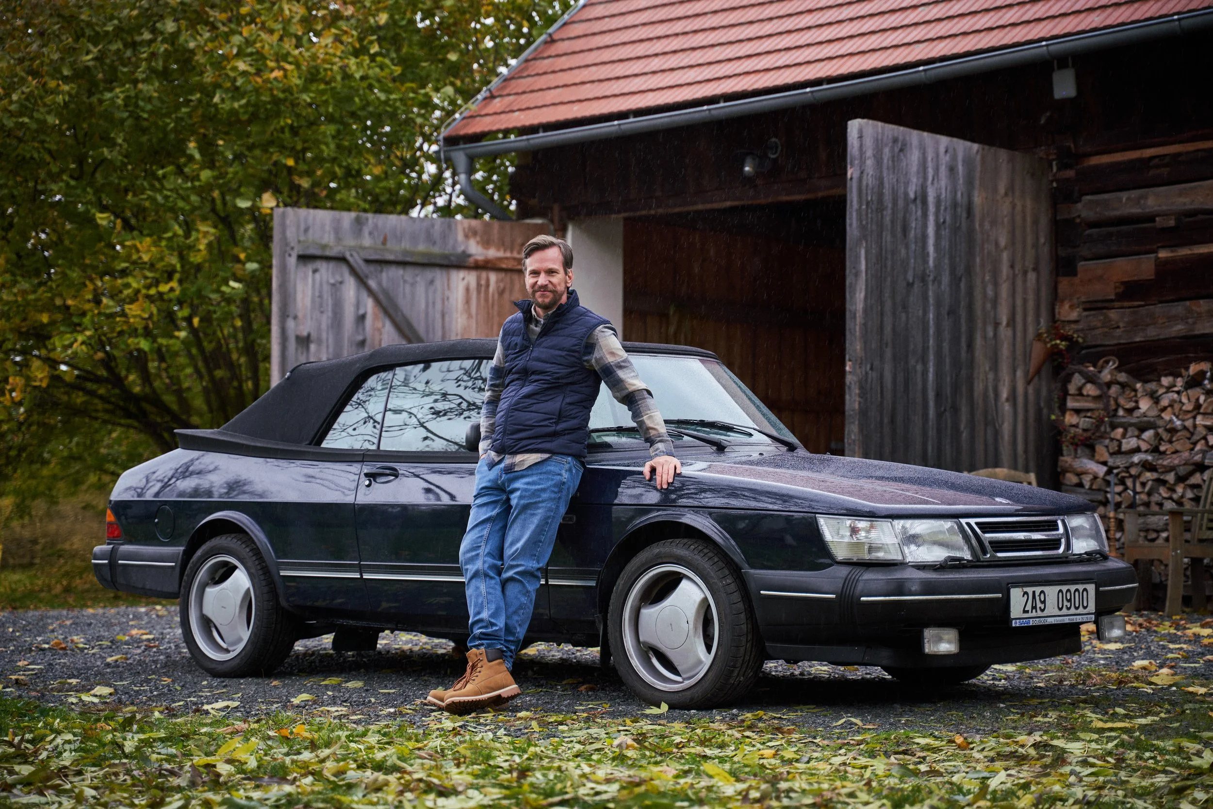 A man leaning on a black Saab convertible car in a driveway, with a wooden shed and a background of trees with falling leaves.