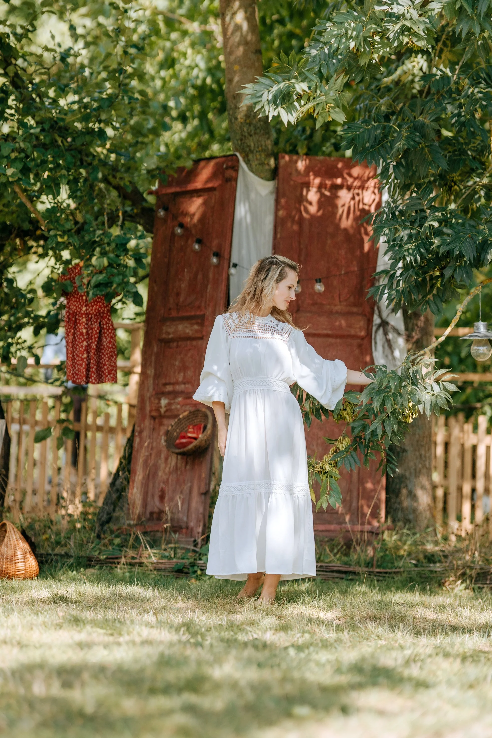 A woman in a long white dress standing outdoors, touching green leaves on a tree, with a rustic red wooden backdrop and string lights hanging, surrounded by greenery.