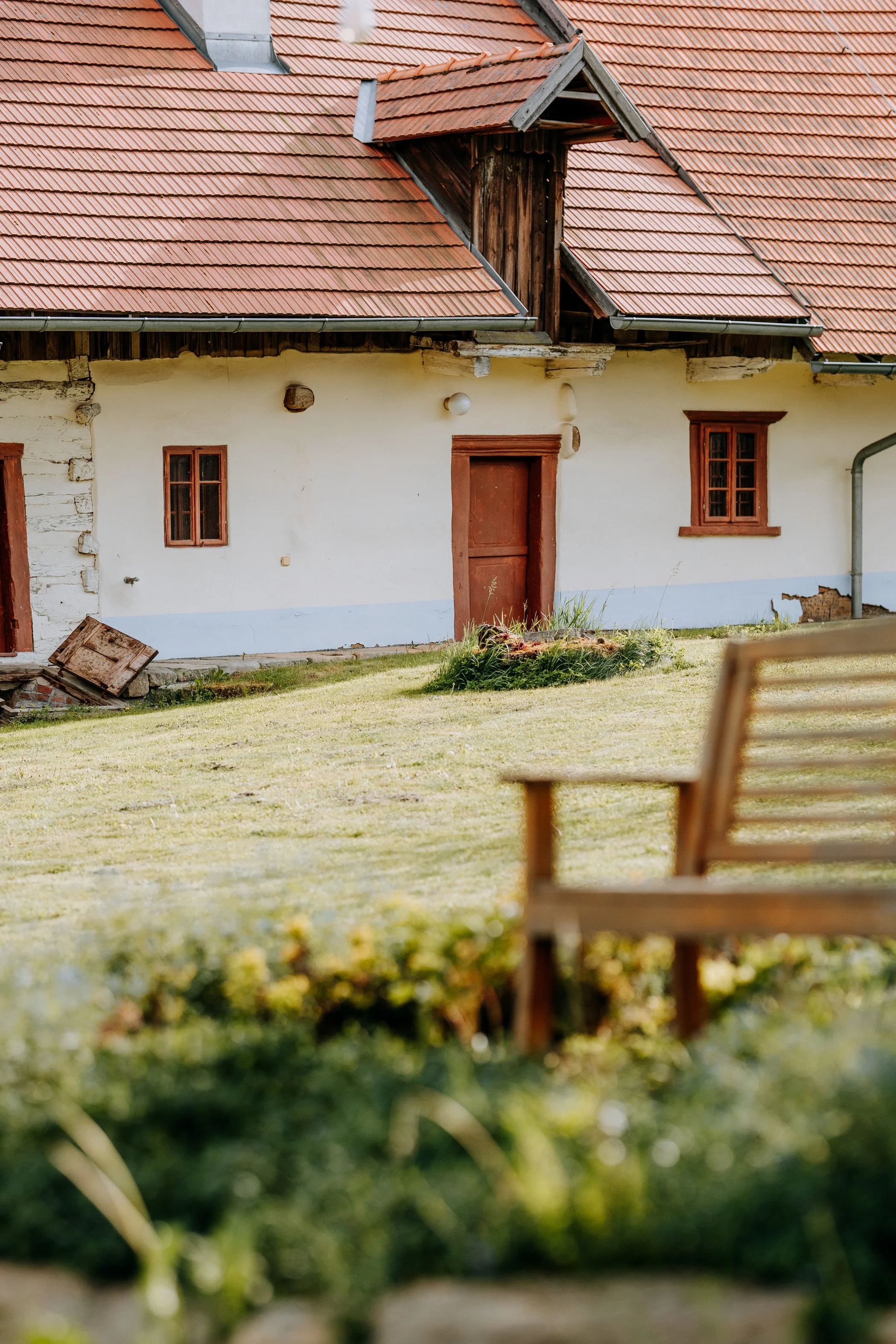 A rustic house with a red tile roof, white walls, and brown window frames, featuring a door and some damaged siding, with a grassy area and a wooden bench in the foreground.