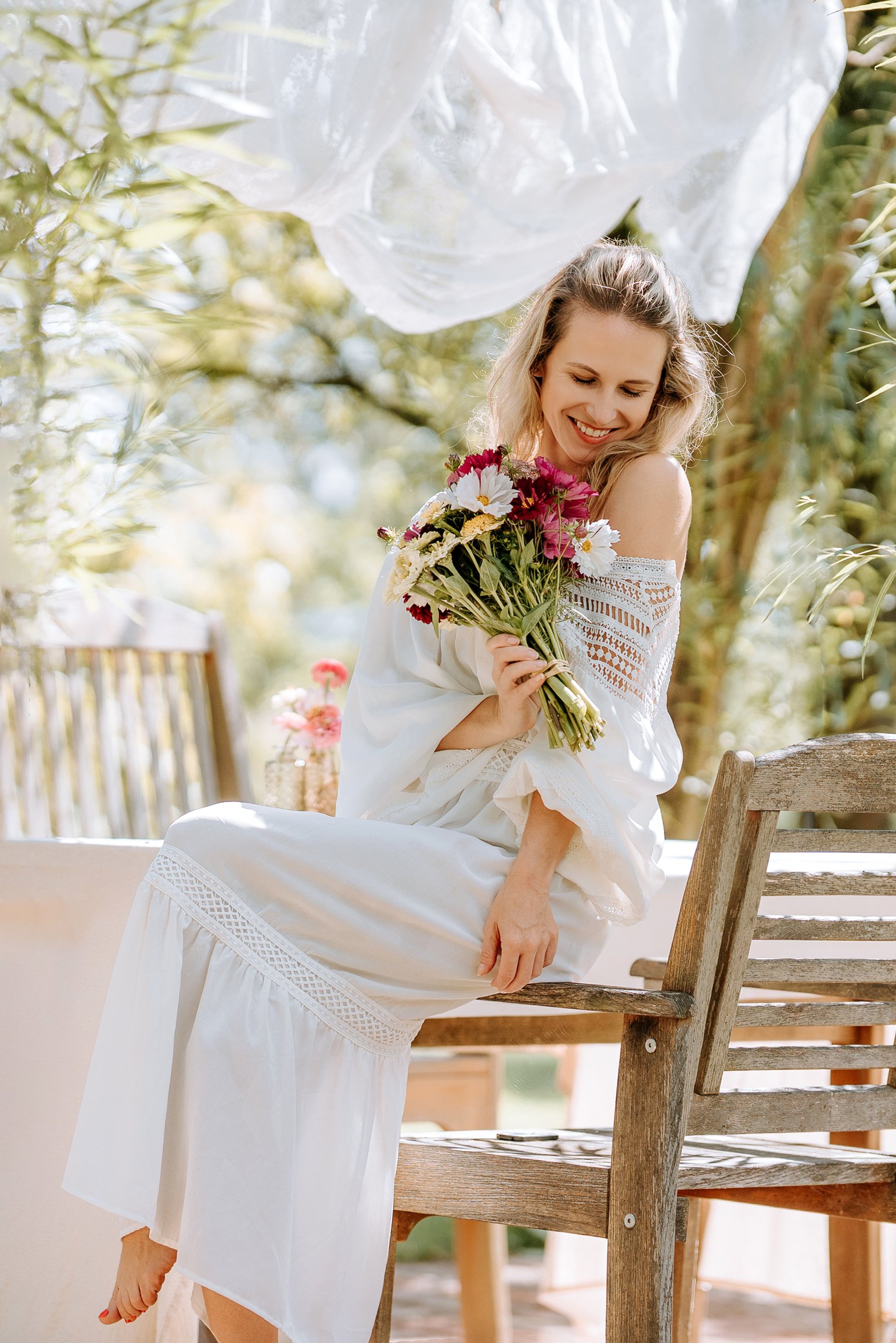 A woman in a white wedding dress sitting on a black motorcycle outdoors on a dirt path with green trees in the background.