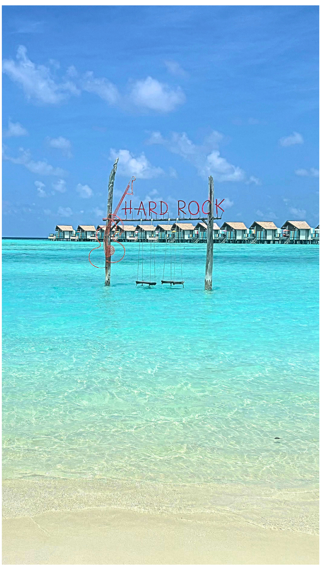 Clear turquoise water at a beach in the Maldives with a wooden swing hanging from two poles. A sign reads 'HARD ROCK' and there are overwater bungalows in the background under blue sky.