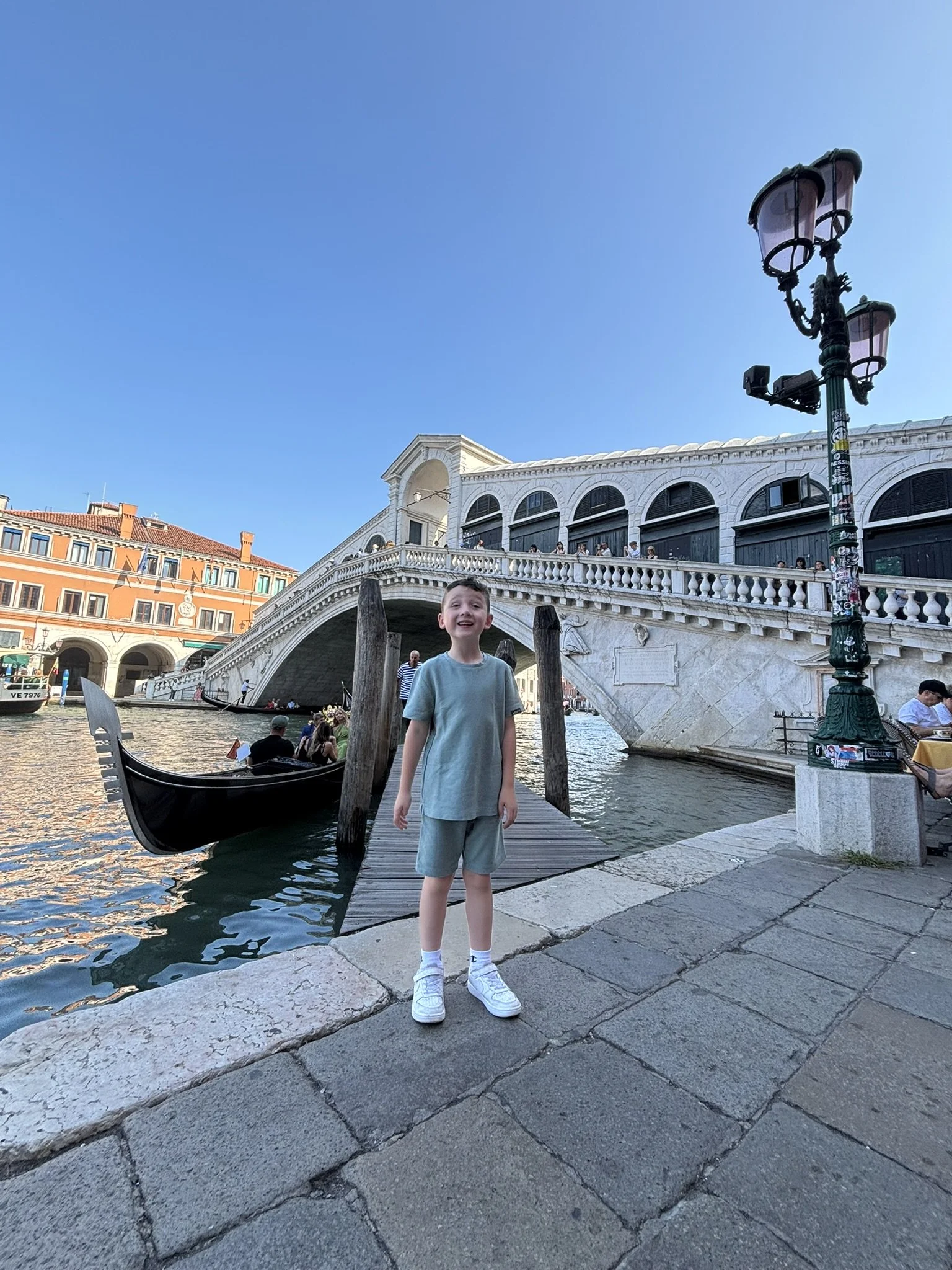 A young boy smiling and standing on a stone walkway beside a canal in Venice, Italy, with the iconic Rialto Bridge in the background and a gondola on the water.