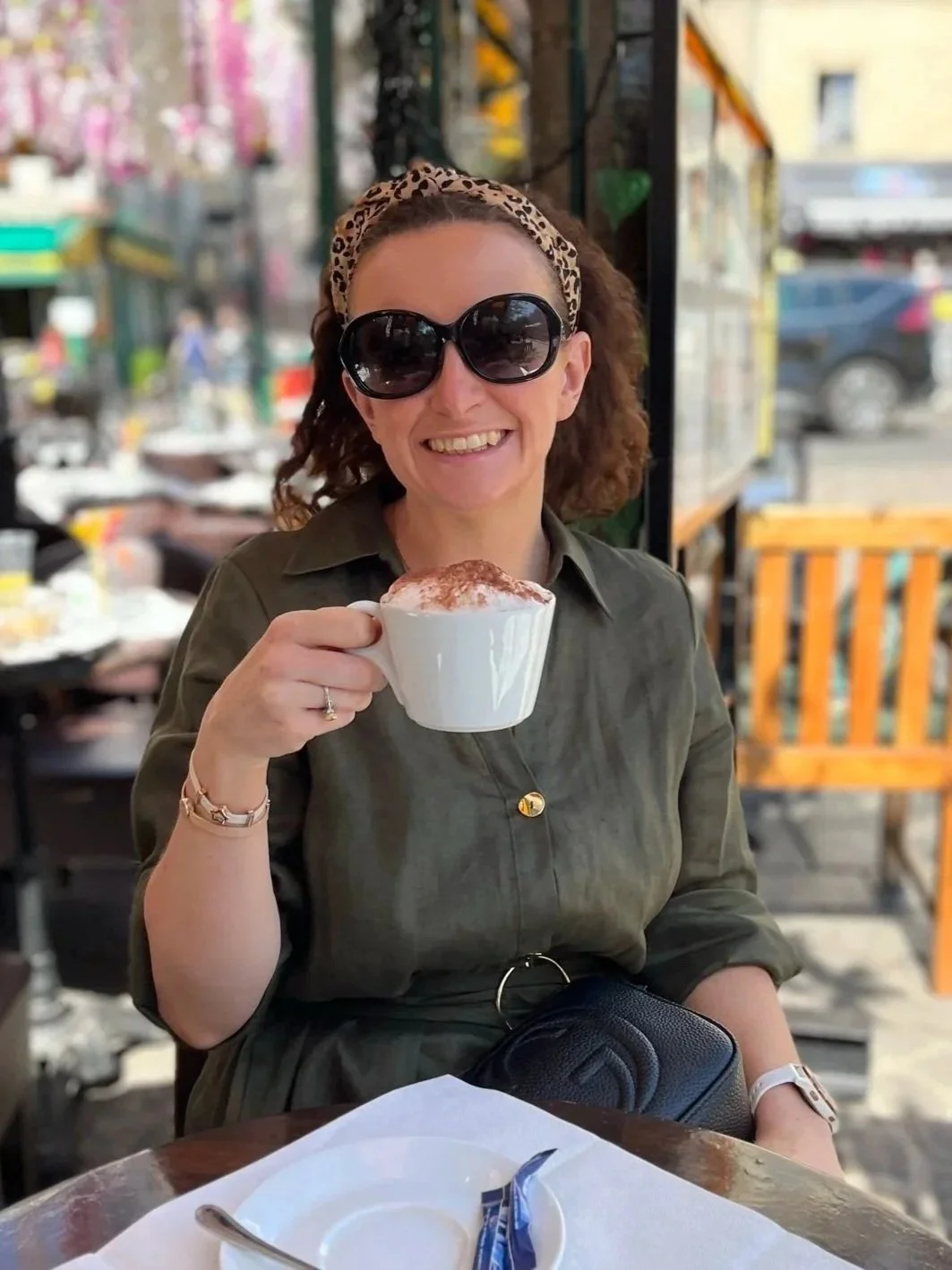 A woman with curly hair, wearing large sunglasses and a leopard print headband, smiling and holding a white mug of cappuccino with cinnamon on top, sitting outdoors at a cafe.
