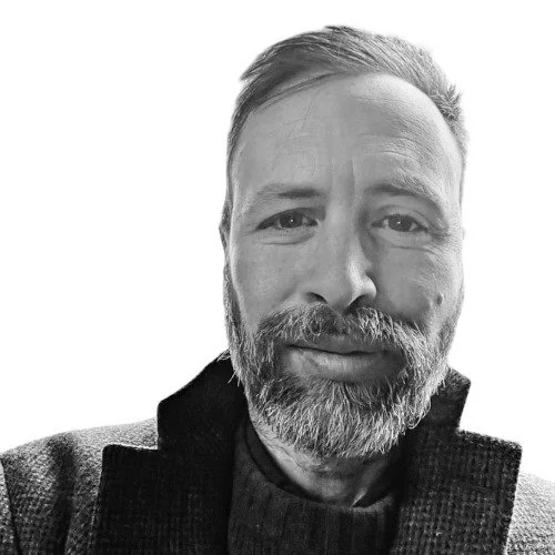 A black and white close-up photo of a smiling man with a beard, wearing a dark jacket and shirt, against a plain white background.