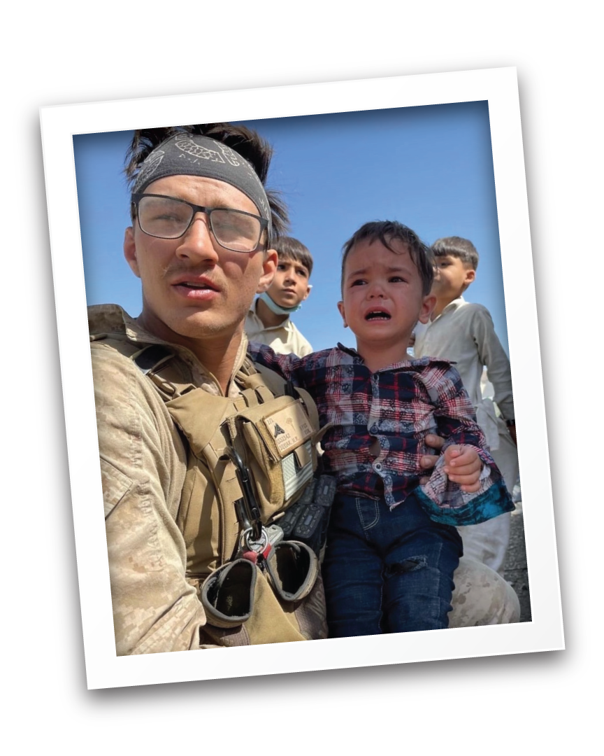 Max in military gear and glasses holding a crying boy in a plaid shirt, with three other children in the background against a blue sky.