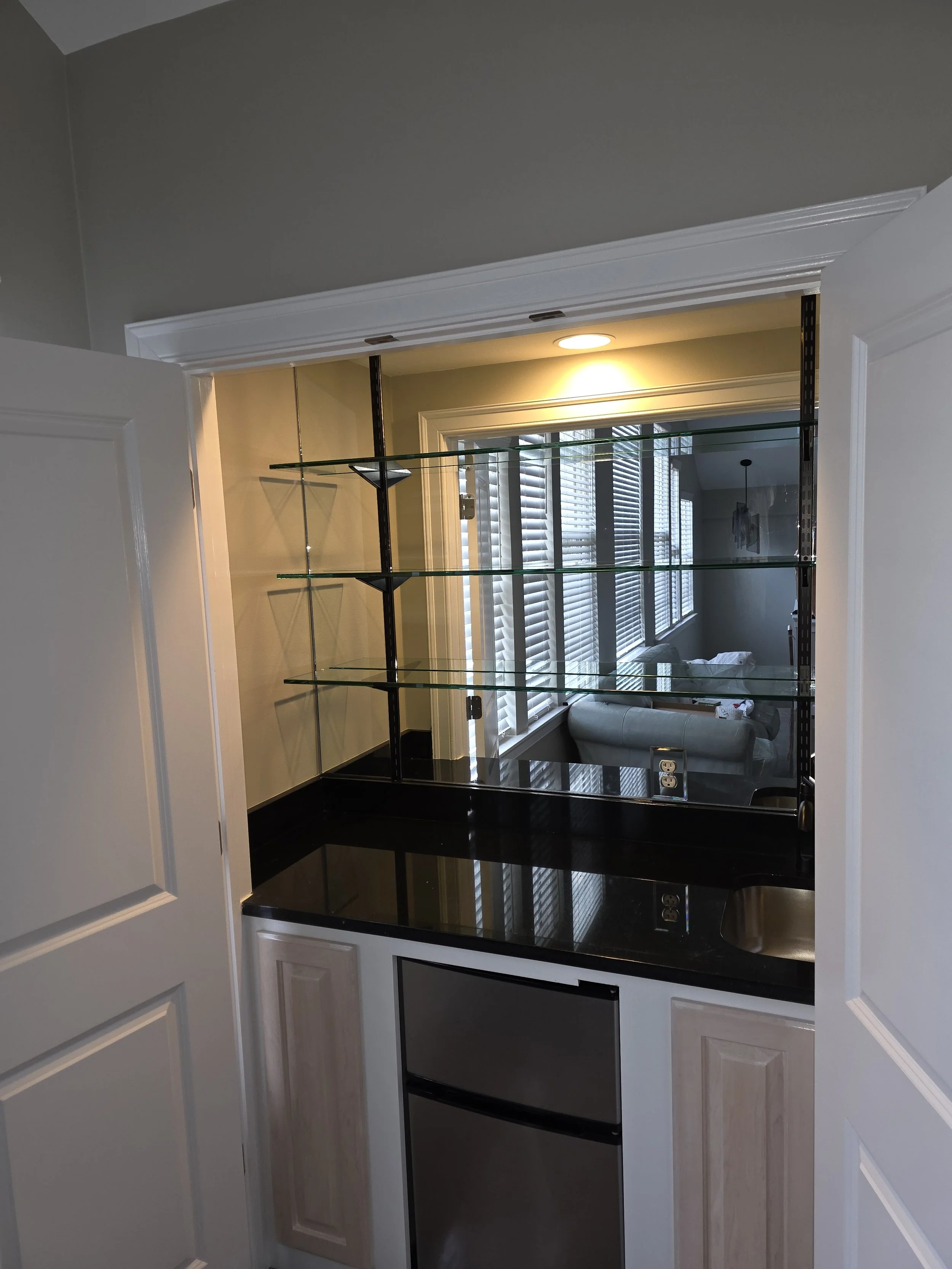 Small kitchen area with open white double doors, black countertop, mini fridge, glass shelves, and a small sink, overlooking a living room with large windows, blinds, and a couch.