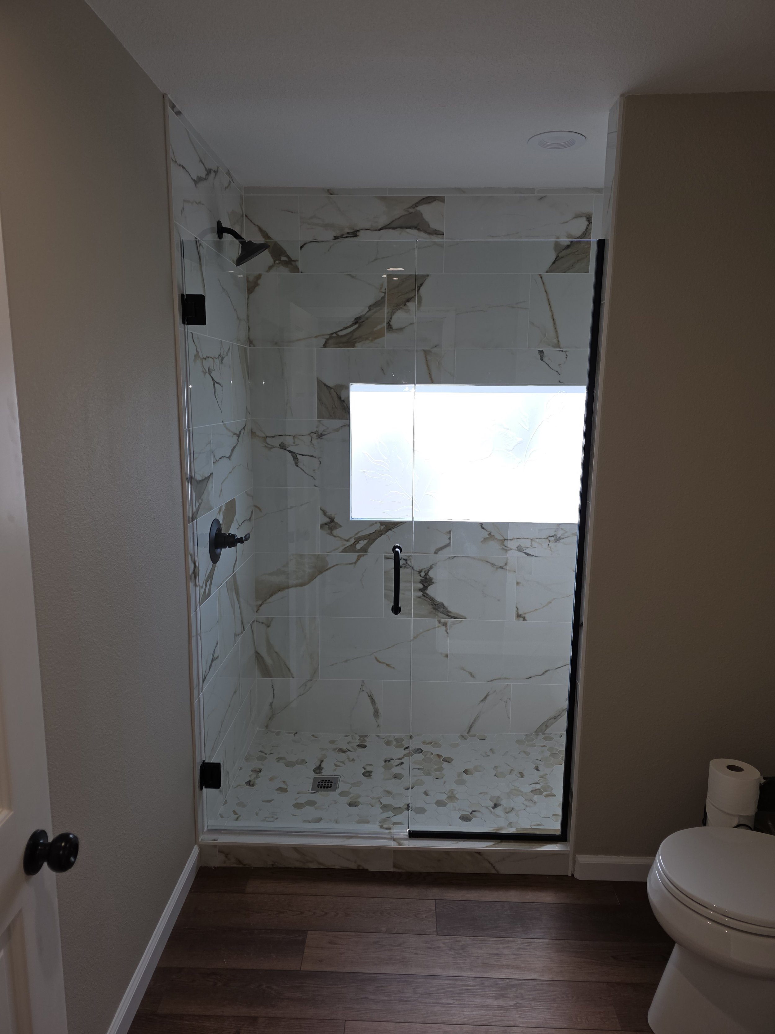 Modern bathroom shower with marble tiles, a glass door, and a window on the back wall. The shower area has a rainfall showerhead, a handheld shower, and a floor with pebble tiles. The bathroom features wooden flooring and a white toilet.