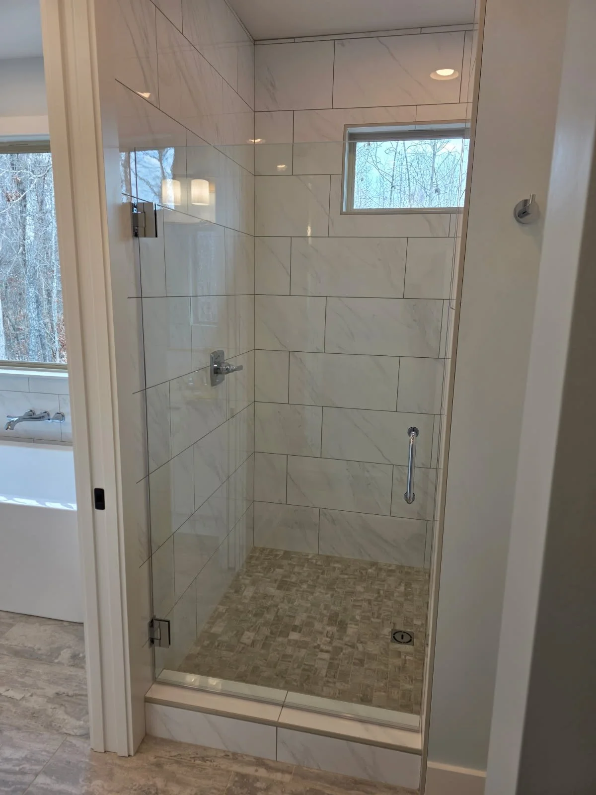 Empty walk-in shower with tiled walls and a small window, next to a bathtub with a window showing outdoor trees.