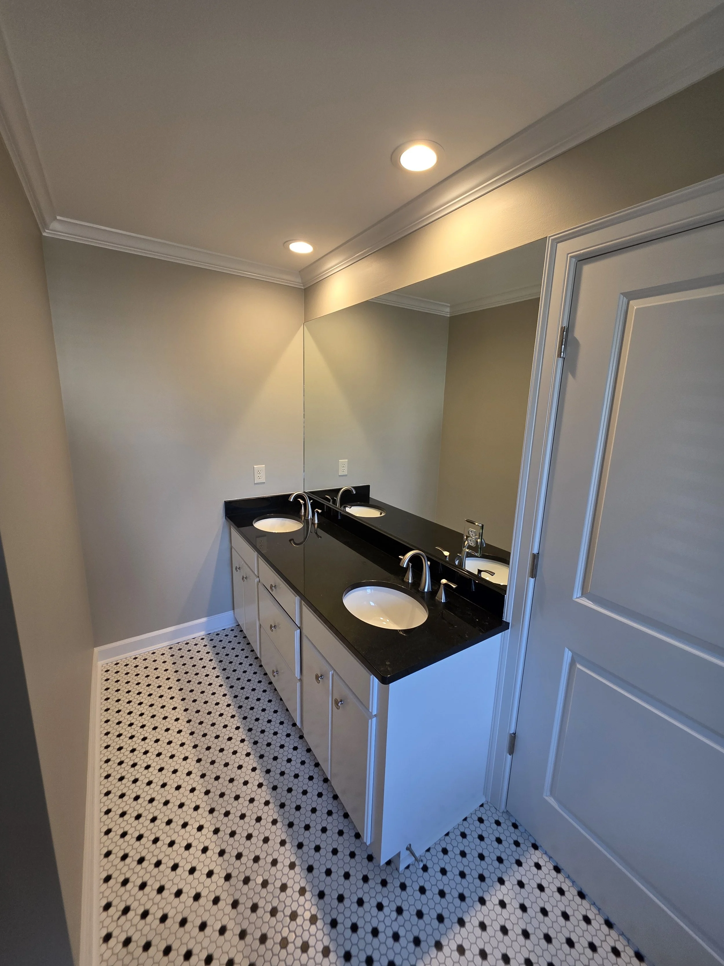 Bathroom with double sink vanity, black countertop, large mirror, beige walls, white cabinetry, and black and white mosaic tile floor.