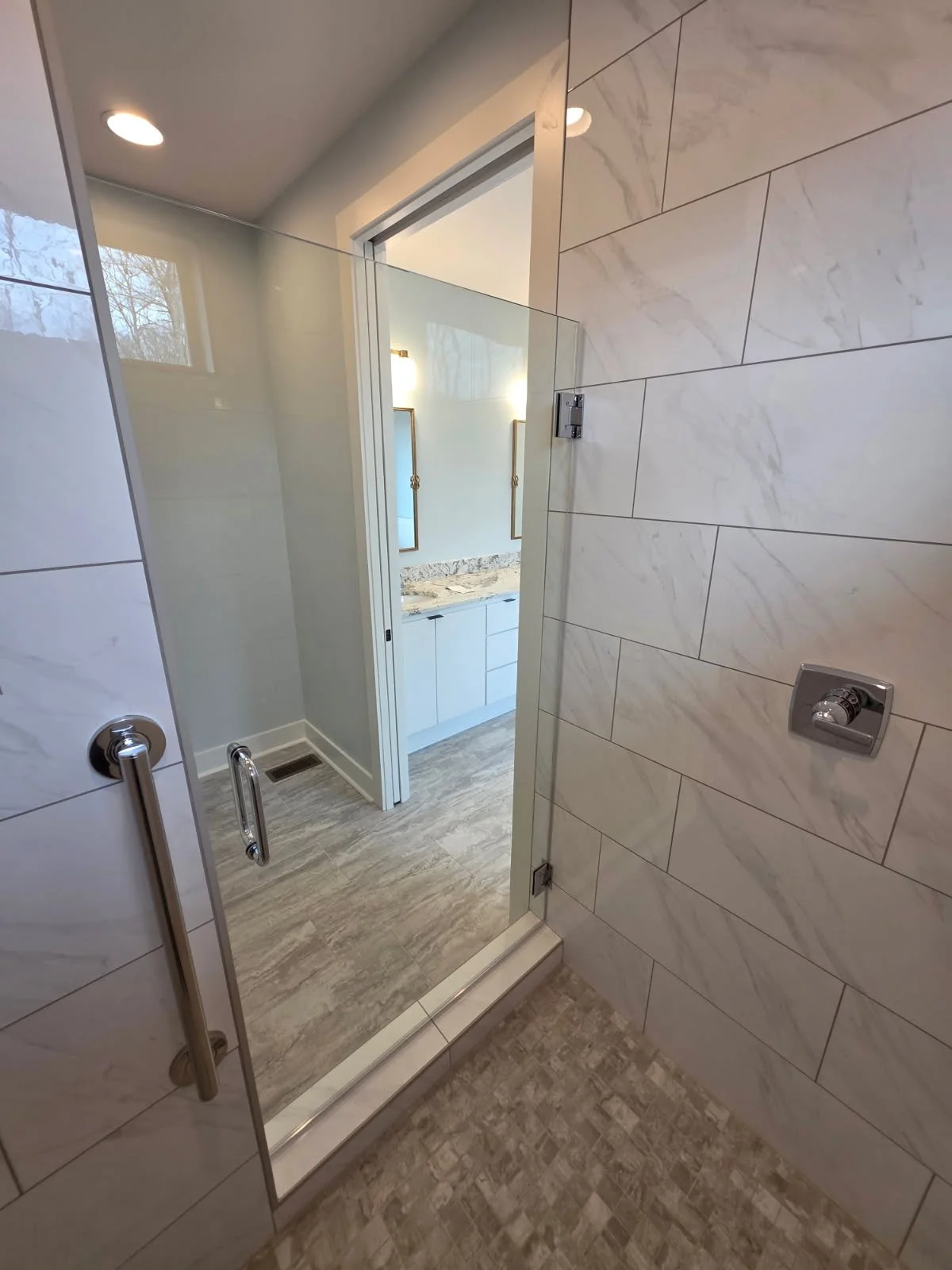 A modern bathroom with a glass shower door, marble wall tiles, and a vanity seen through the open door.