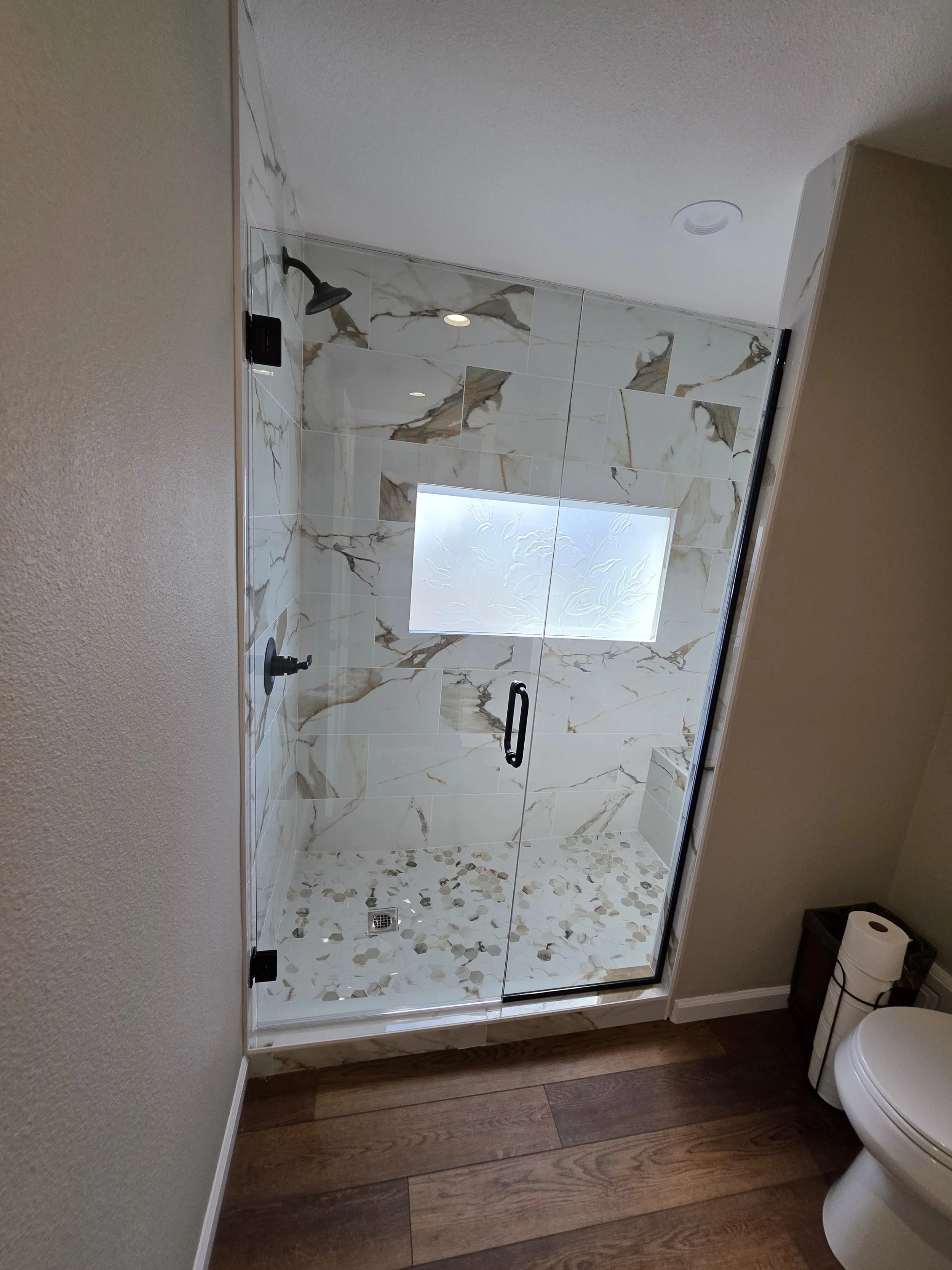 Modern walk-in shower with glass door, marble tiles, and a frosted window, adjacent to a toilet and wooden floor.