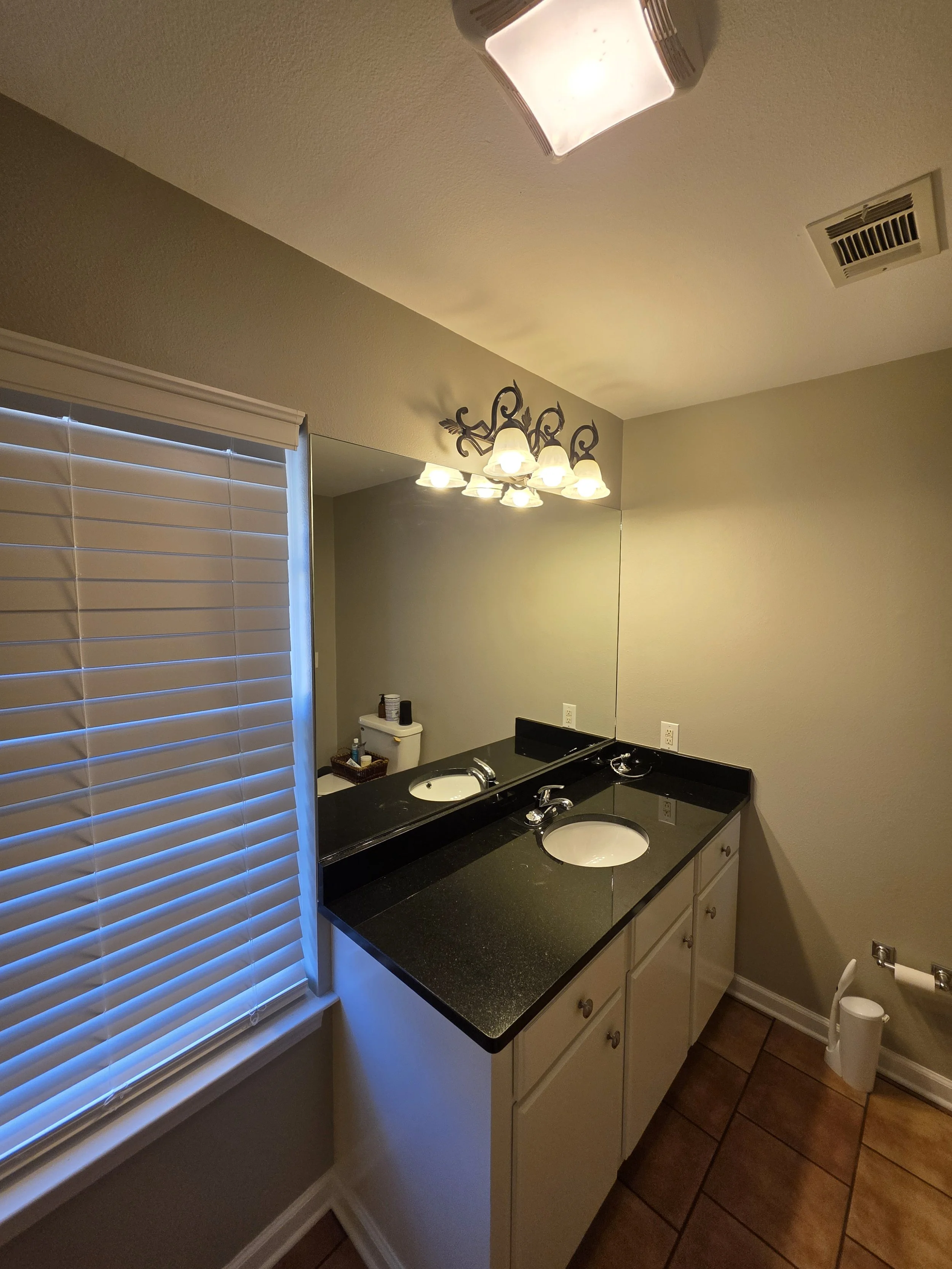 Bathroom sink with black countertop, large mirror, vanity lights, window with blinds, and a toilet in the background.