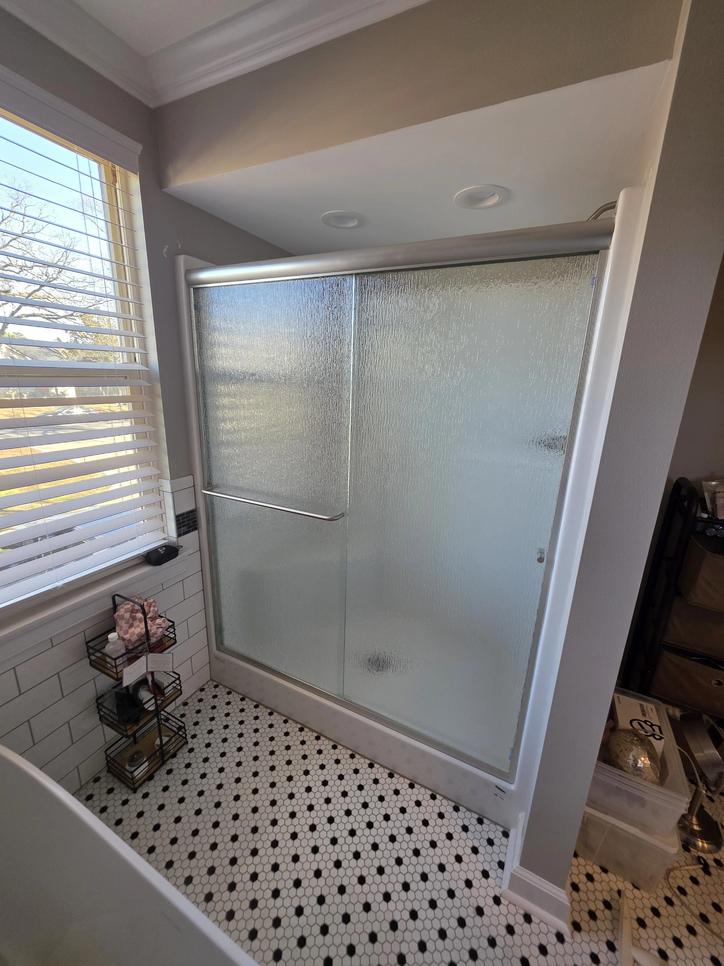 Bathroom shower with frosted glass door, black and white hexagonal tiled floor, white subway tile walls, window with blinds, and storage rack.