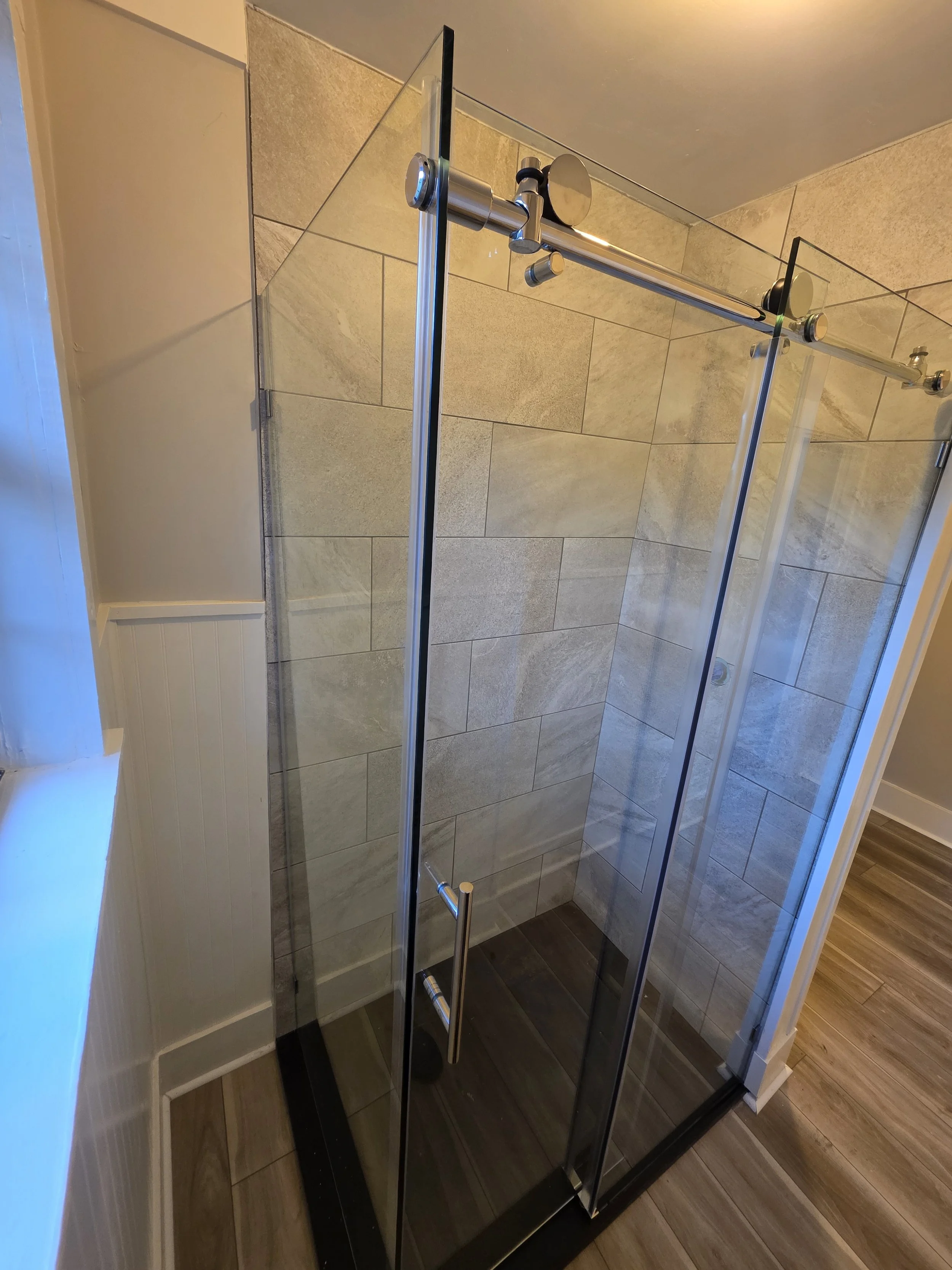 Glass shower enclosure with metal hardware and a handle, tiled walls, and wood-look flooring in a bathroom.