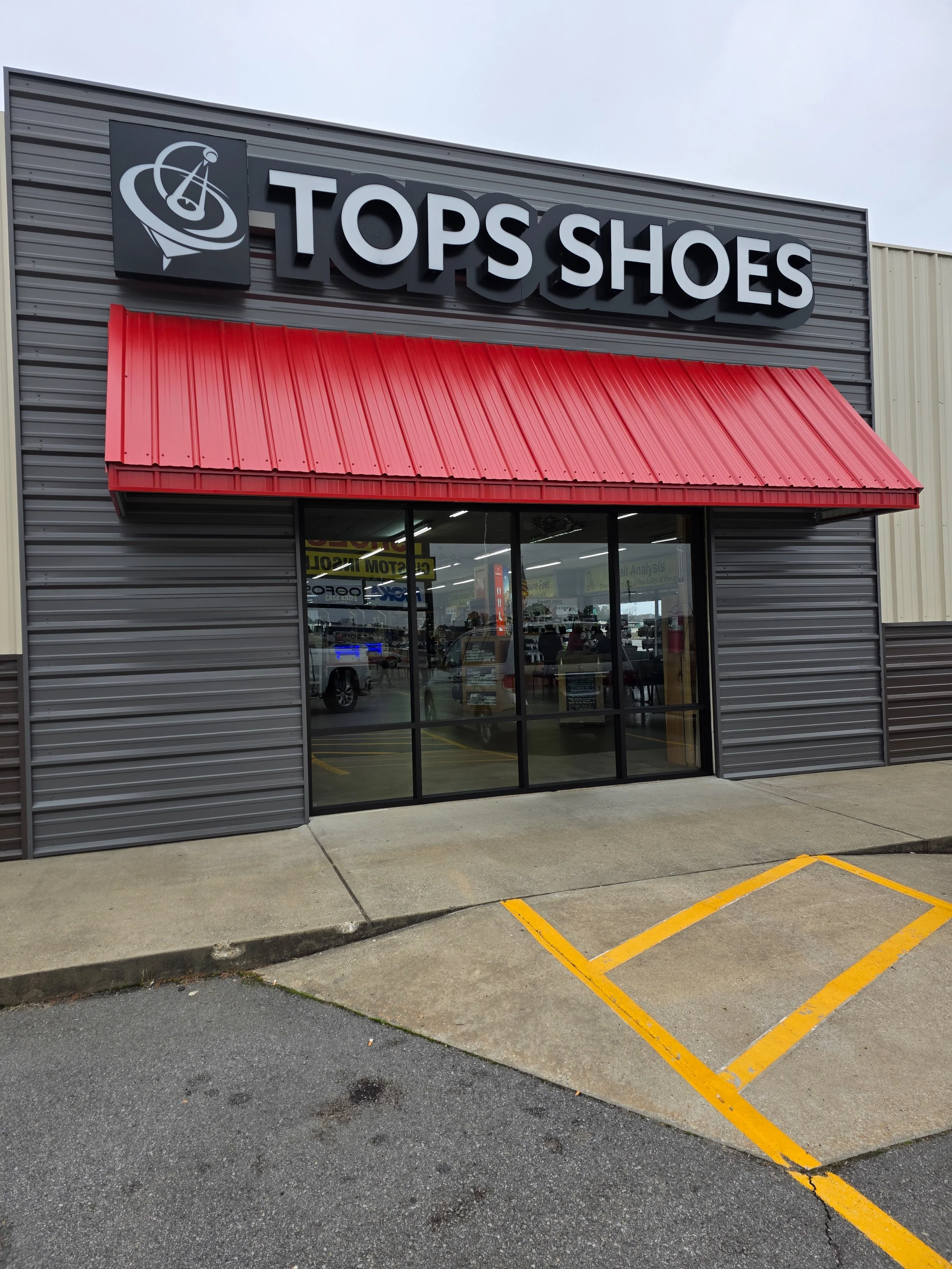 Exterior of a shoe store named 'TOPS SHOES' with a black sign and white lettering, and a red metal awning over glass entrance doors. Parking space with yellow lines in front.