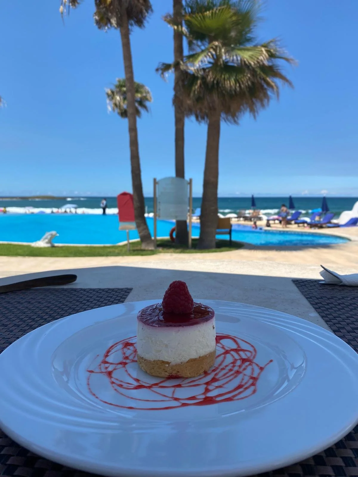 Un dessert en forme de tartelette avec une framboise sur le dessus, présenté sur une assiette blanche avec un filet de sauce rouge, à l'extérieur près d'une piscine avec des palmiers et des parasols.