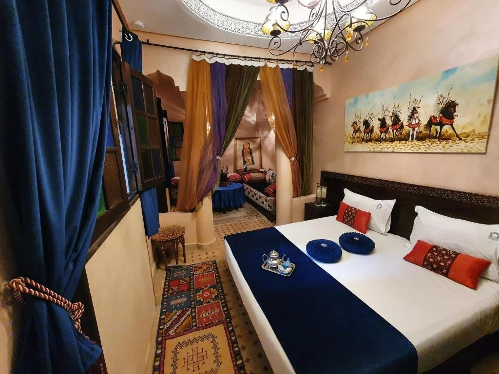Chambre décorée avec draperies colorées, lit avec coussins rouges et bleus, tableau représentant des chameaux dans le désert, et un coin salon avec tapis berbère.