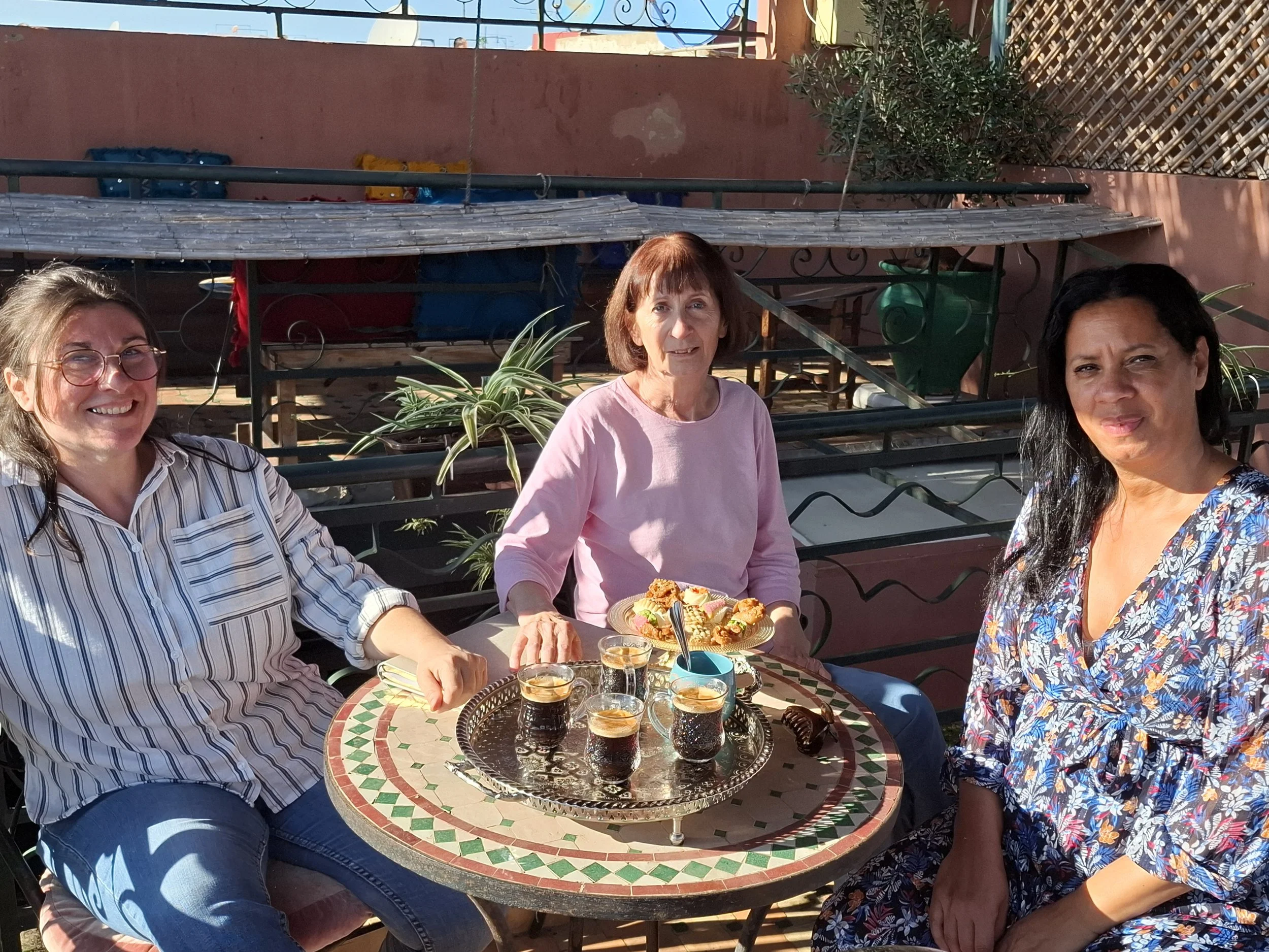 Trois femmes assises autour d'une table avec des cafés et des pâtisseries, profitant du soleil sur une terrasse extérieure.