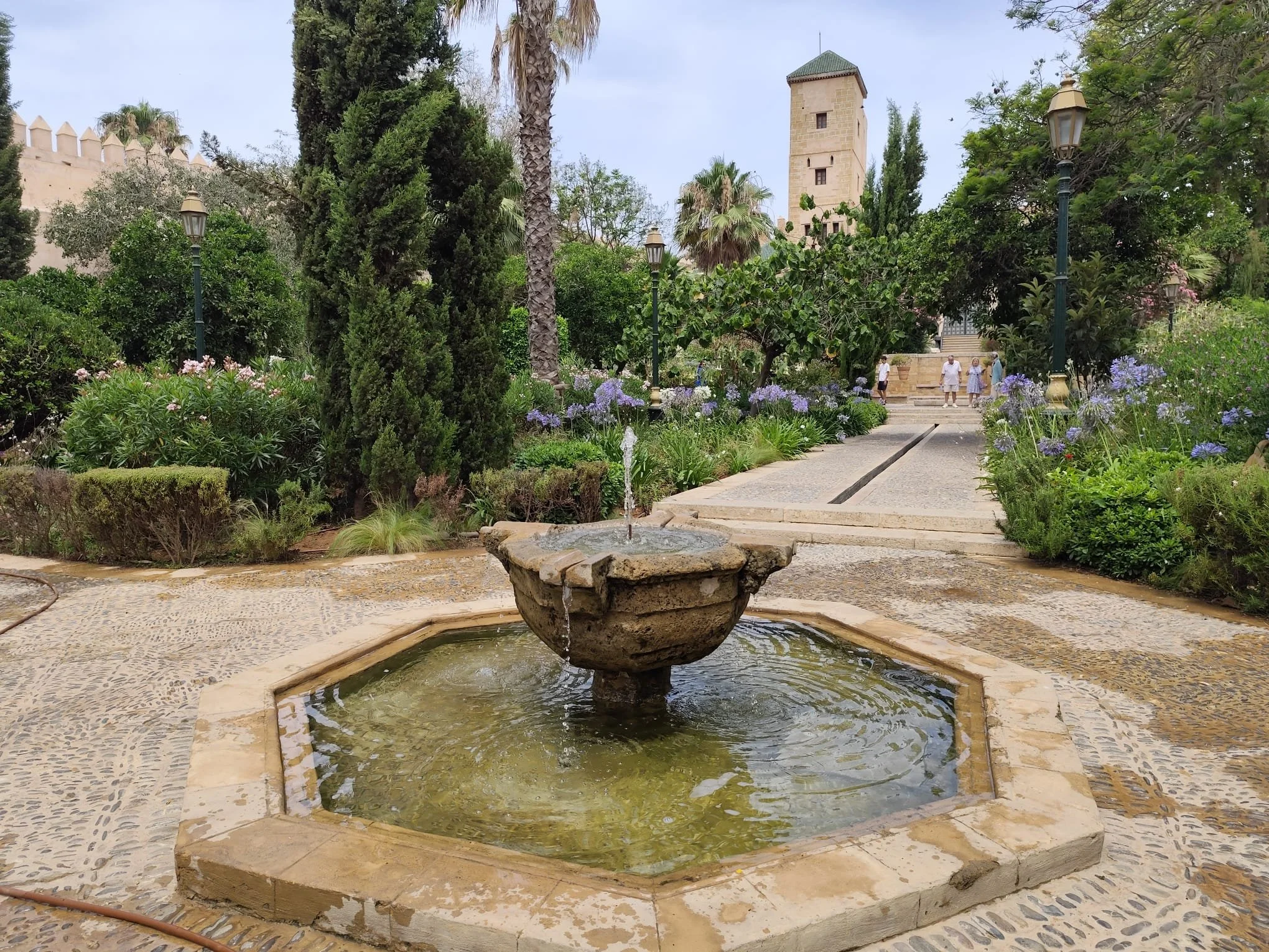 Une fontaine en pierre au centre d'une cour pavée, entourée de végétation, avec un chemin menant à une tour en pierre au fond, dans un jardin verdoyant.