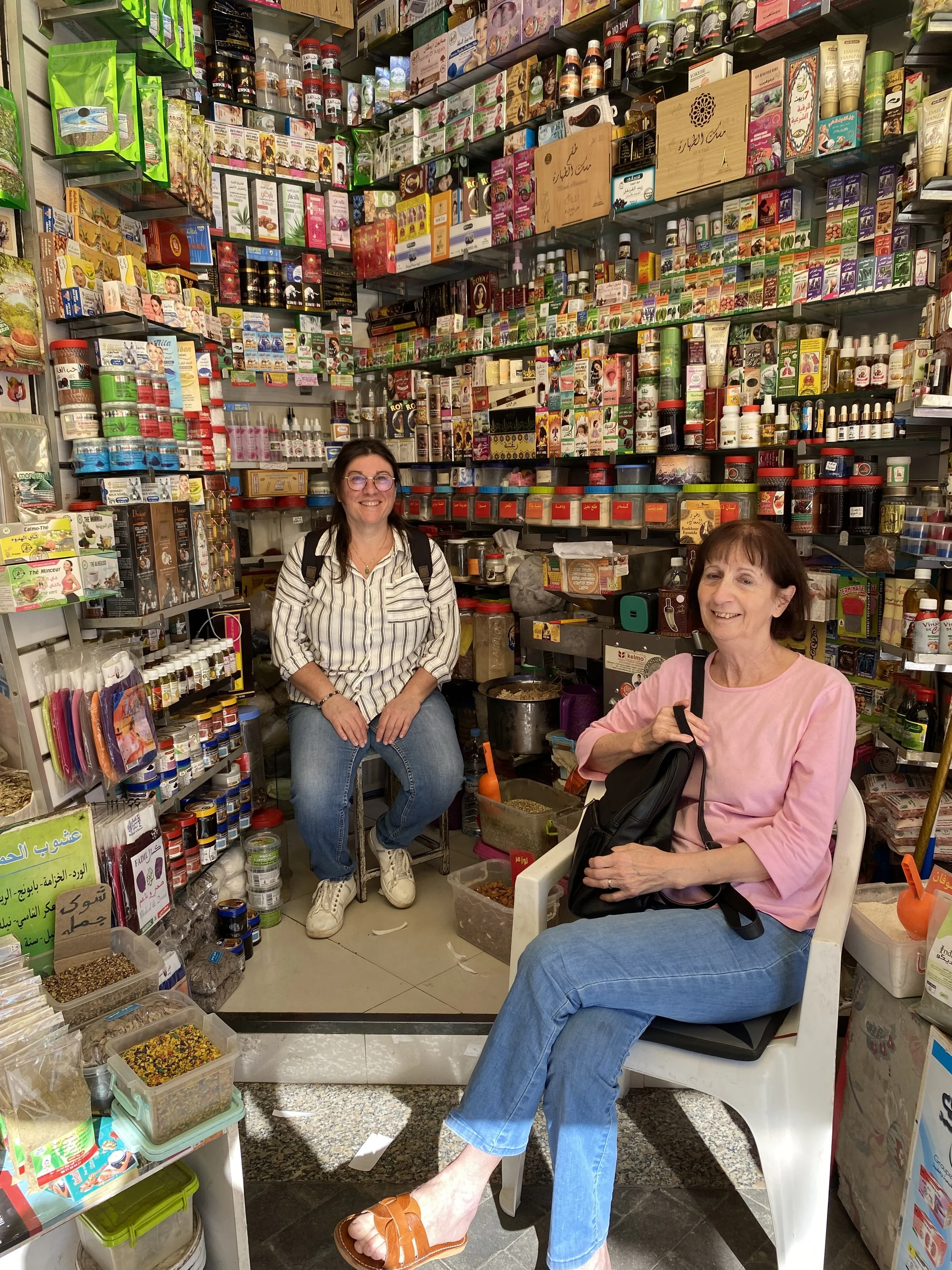Deux femmes dans une boutique proposant des produits divers, notamment des épices, des herbes, et des produits de beauté, avec beaucoup de bocaux, de boîtes et d'emballages colorés