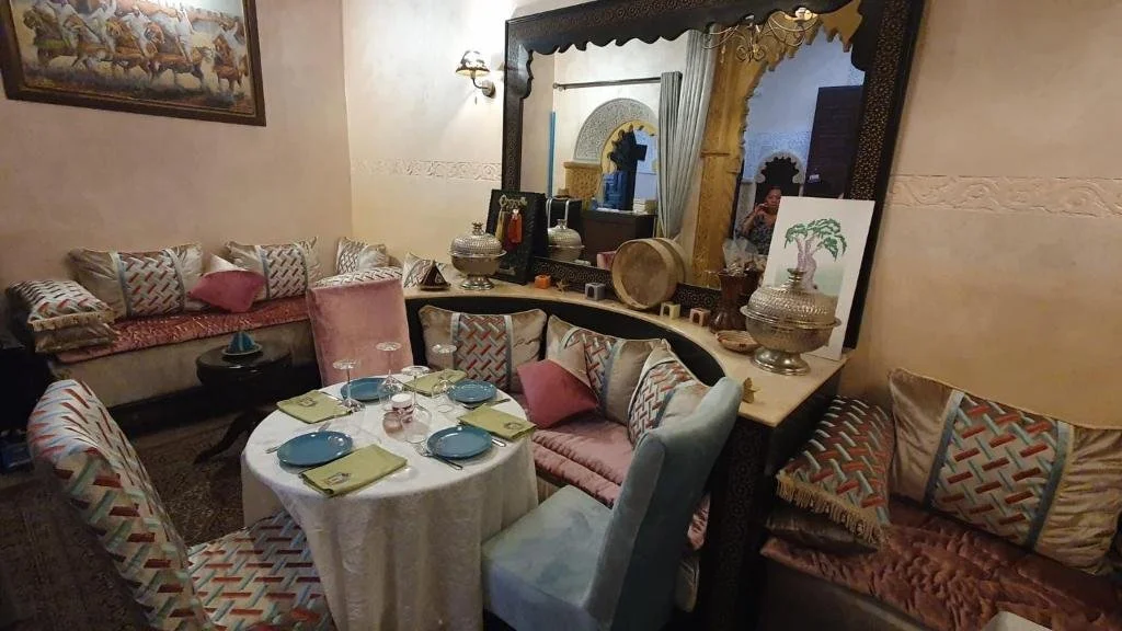 Une salle à manger décorée dans un style marocain avec un miroir orné, un tableau, et un mobilier avec des coussins colorés autour de la table dressée pour un repas.