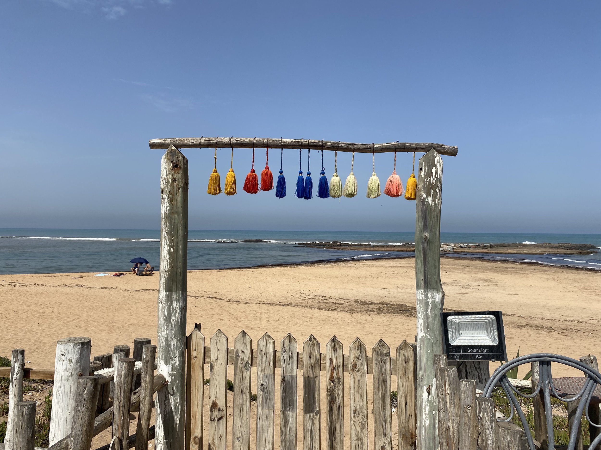 Une plage avec un cadre en bois avec des pompons colorés suspendus, la mer en arrière-plan, ciel clair, quelques personnes sous un parasol.