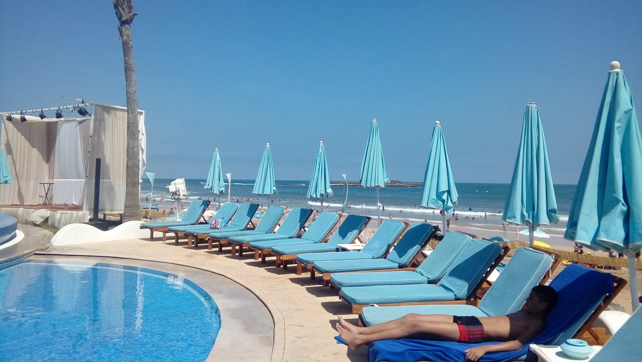 Chaises longues bleues sous parasols au bord de la mer, plage en arrière-plan, personne allongée sur une chaise longue, mer et ciel bleus.