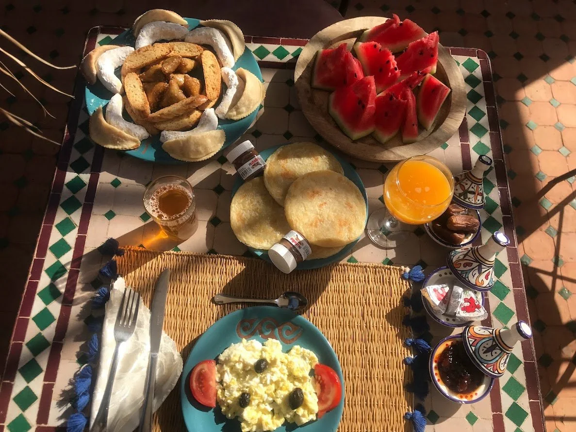 Un repas avec des plats et des boissons traditionnels, comprenant une salade de pommes de terre, des tranches de pastèque, des falafels, des morceaux de pain avec une sauce, un verre de jus d'orange, un verre de bière et plusieurs petits plats avec des sauces ou condiments, le tout sur une table avec une nappe en rotin et un motif géométrique