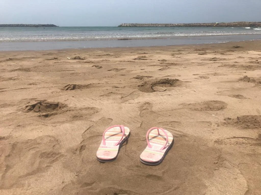 Une paire de tongs roses et blanches sur une plage de sable, avec la mer et le ciel gris en arrière-plan.