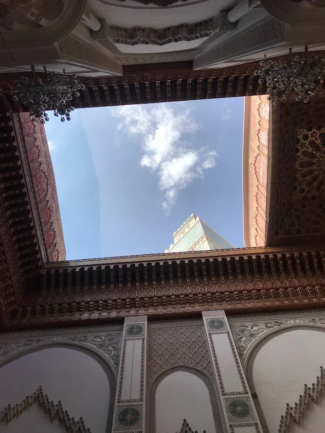 Vue du ciel depuis une cour intérieure ornée de motifs architecturaux marocains traditionnels, avec un bâtiment moderne visible au loin contre un ciel bleu avec quelques nuages.