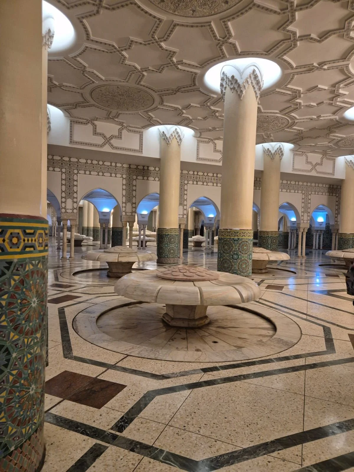 Intérieur d'une salle ornée de colonnes décoratives, plafonds avec motifs traditionnels, et fontaines rondes en marbre.