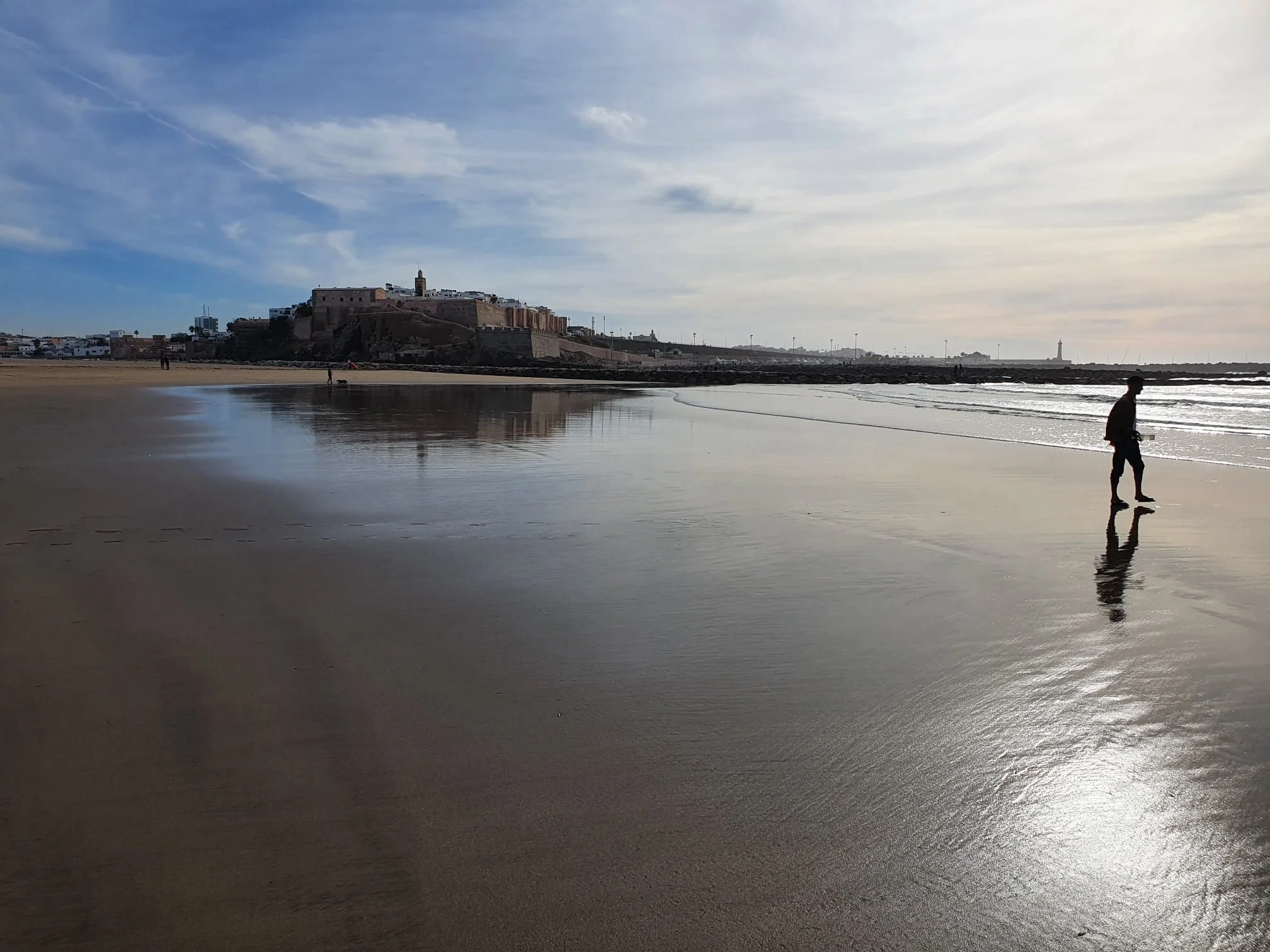 Une plage avec un bâtiment historique en arrière-plan et une personne marchant le long de la côte, avec des reflets dans l'eau calme.