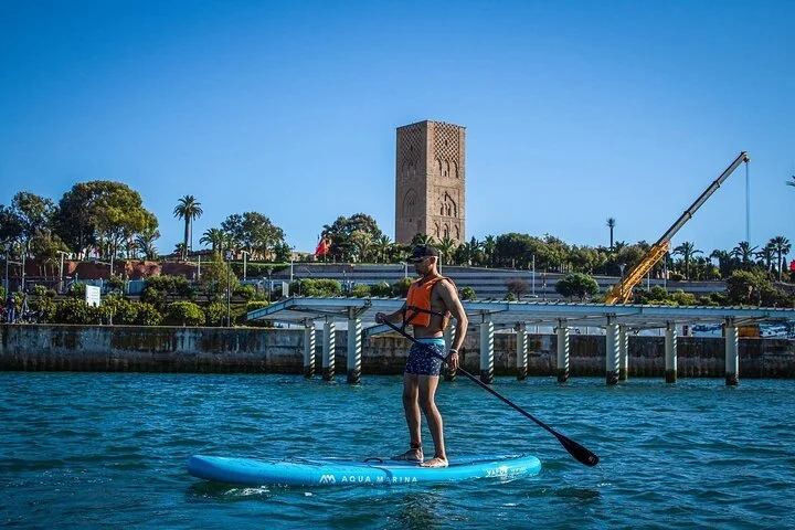 Hommes faisant du paddleboard dans une marina, avec un bâtiment et des palmiers en arrière-plan.