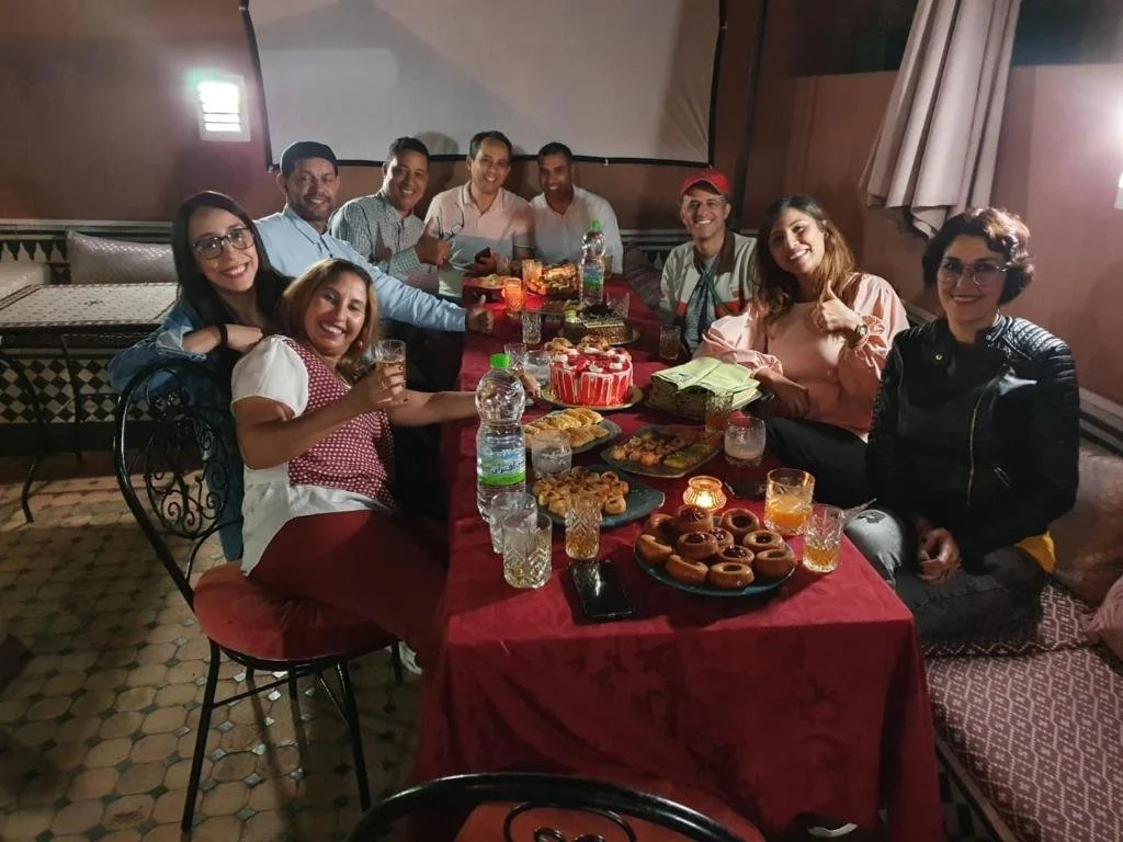 Groupe de personnes assises autour d'une table avec gâteau, boissons, et desserts lors d'une célébration.
