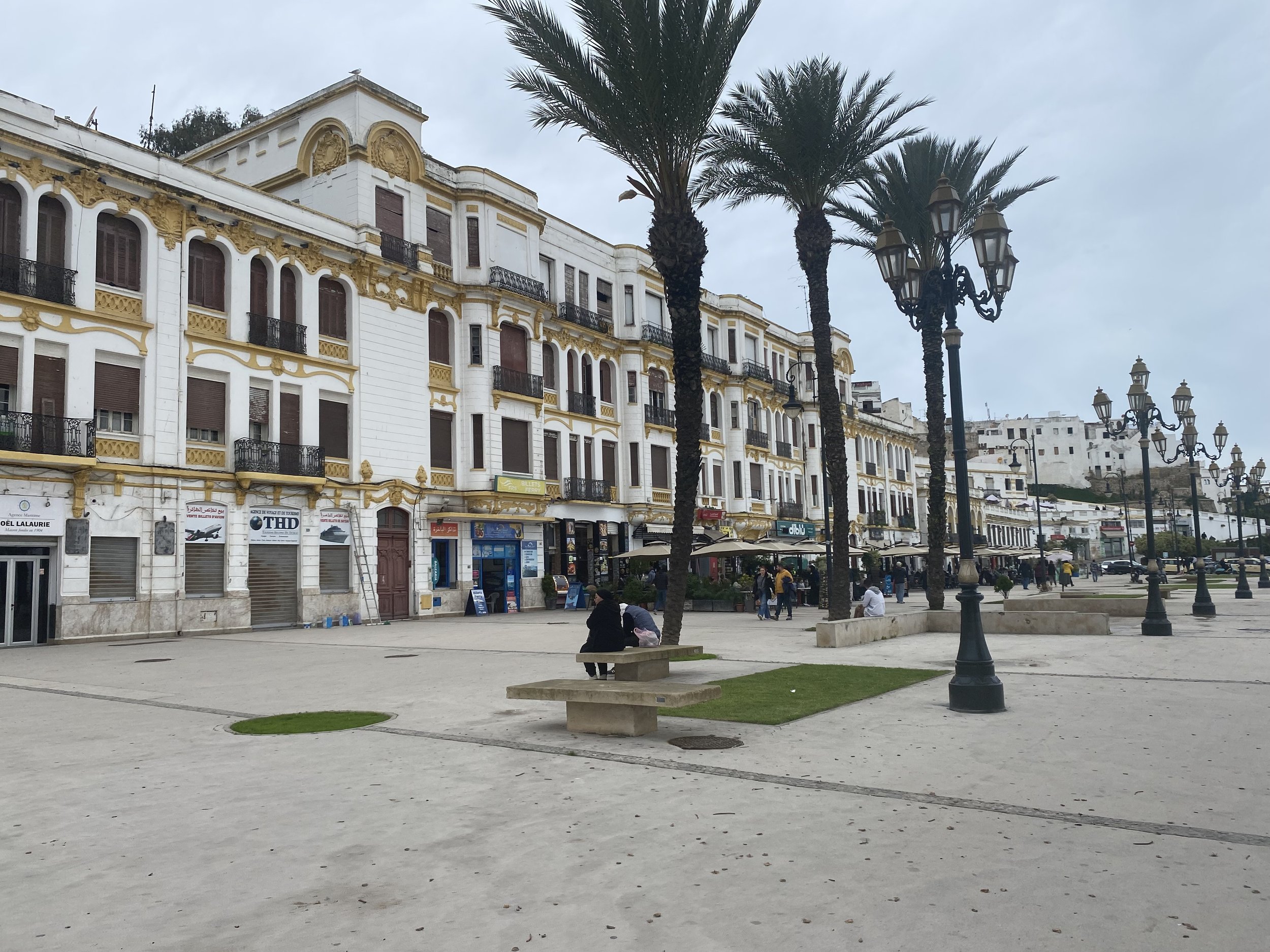 Une place publique bordée d'immeubles blancs avec des balcons ornés de détails dorés, entourée de palmiers et de lampadaires noirs. Quelques personnes assises ou marchant.