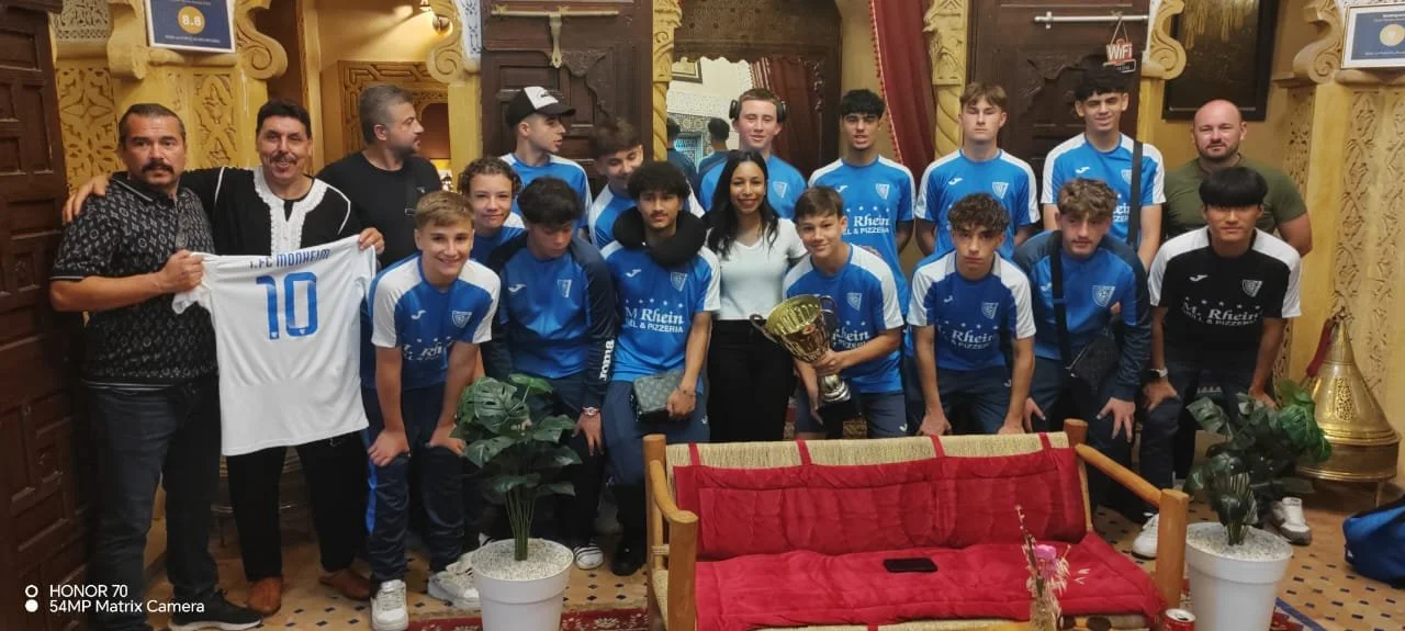 Un groupe de jeunes joueurs de football, accompagnés de leurs entraîneurs et d'une femme, pose pour une photo en intérieur. Certains tiennent un trophée, et l'un d'eux tient un maillot de football blanc avec le numéro 10.