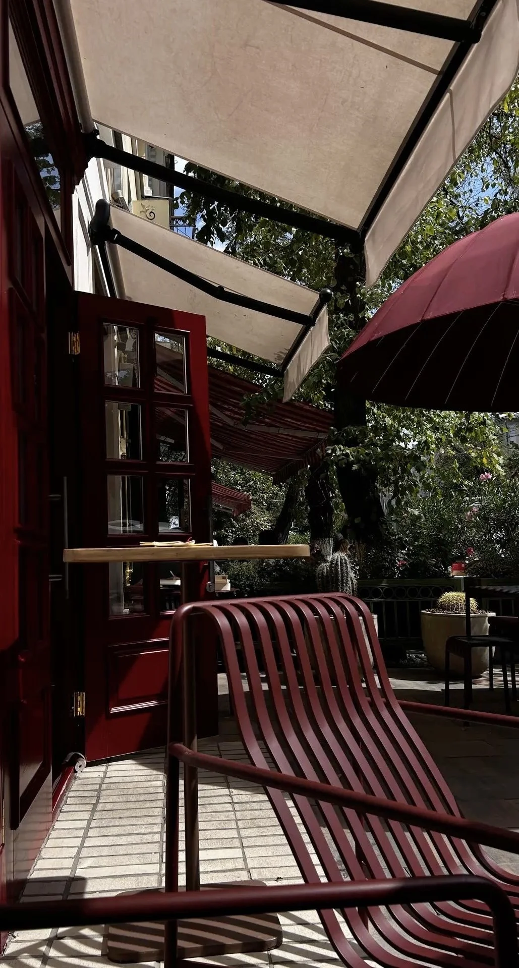 Outdoor patio area with red chairs, potted cactus plants, and tree with hanging plants, shaded by white and red awnings and a pink umbrella.