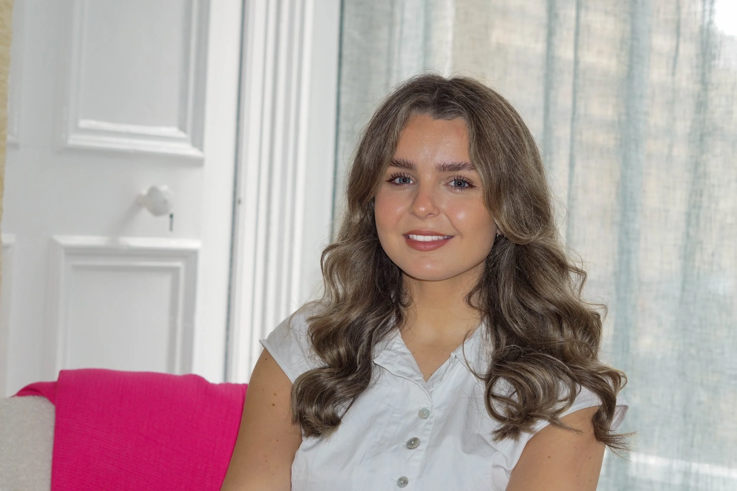 A young woman with wavy brown hair and blue eyes smiling while sitting on a pink chair in front of a window with a light-colored curtain.