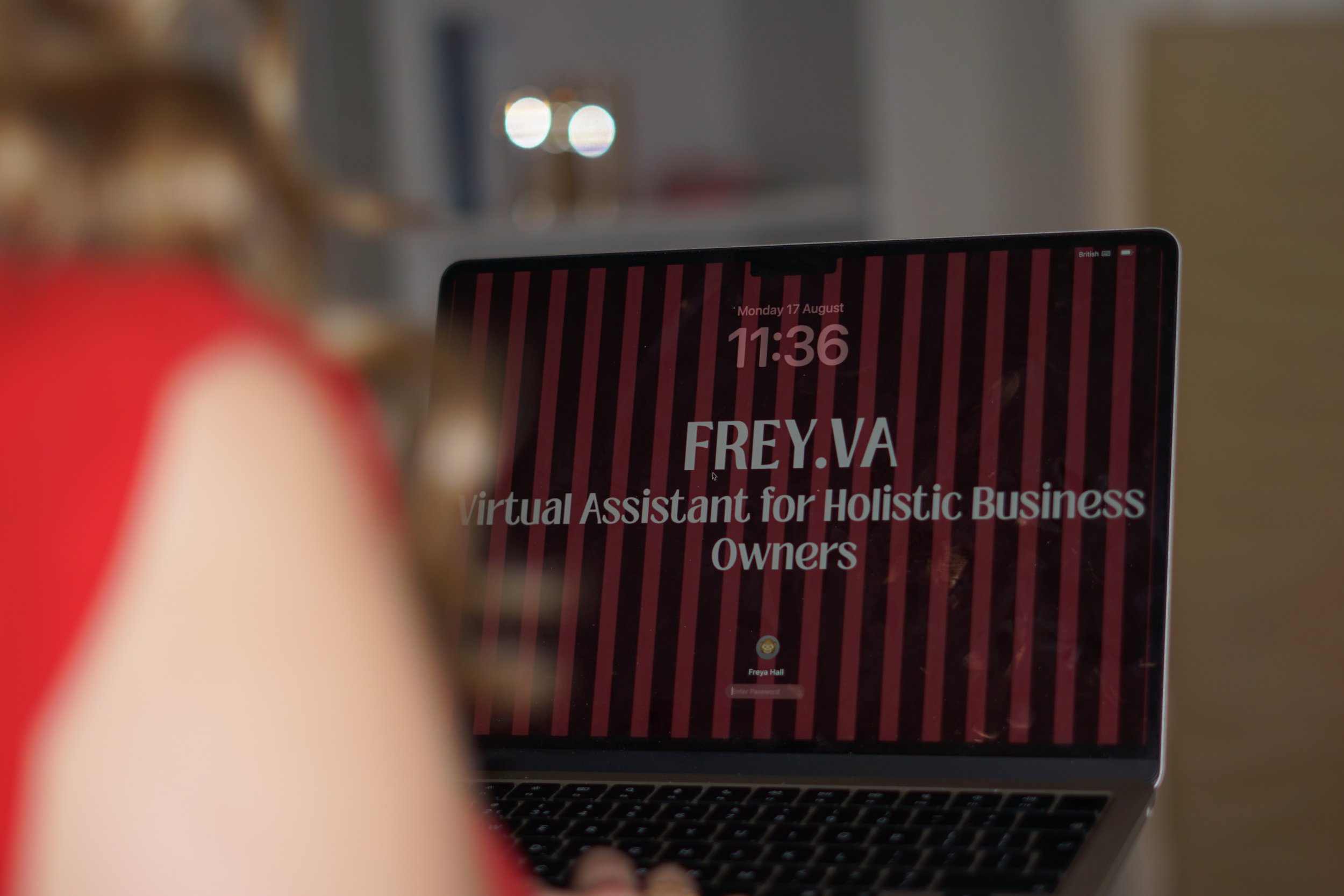 A person holding a smartphone displaying a webpage with the headline "FREY.VA" and the subheading "Virtual Assistant for Holistic Business Owners". The background of the webpage has pink and red vertical stripes, and the date/time on the phone is Monday, 17 August, 11:36.