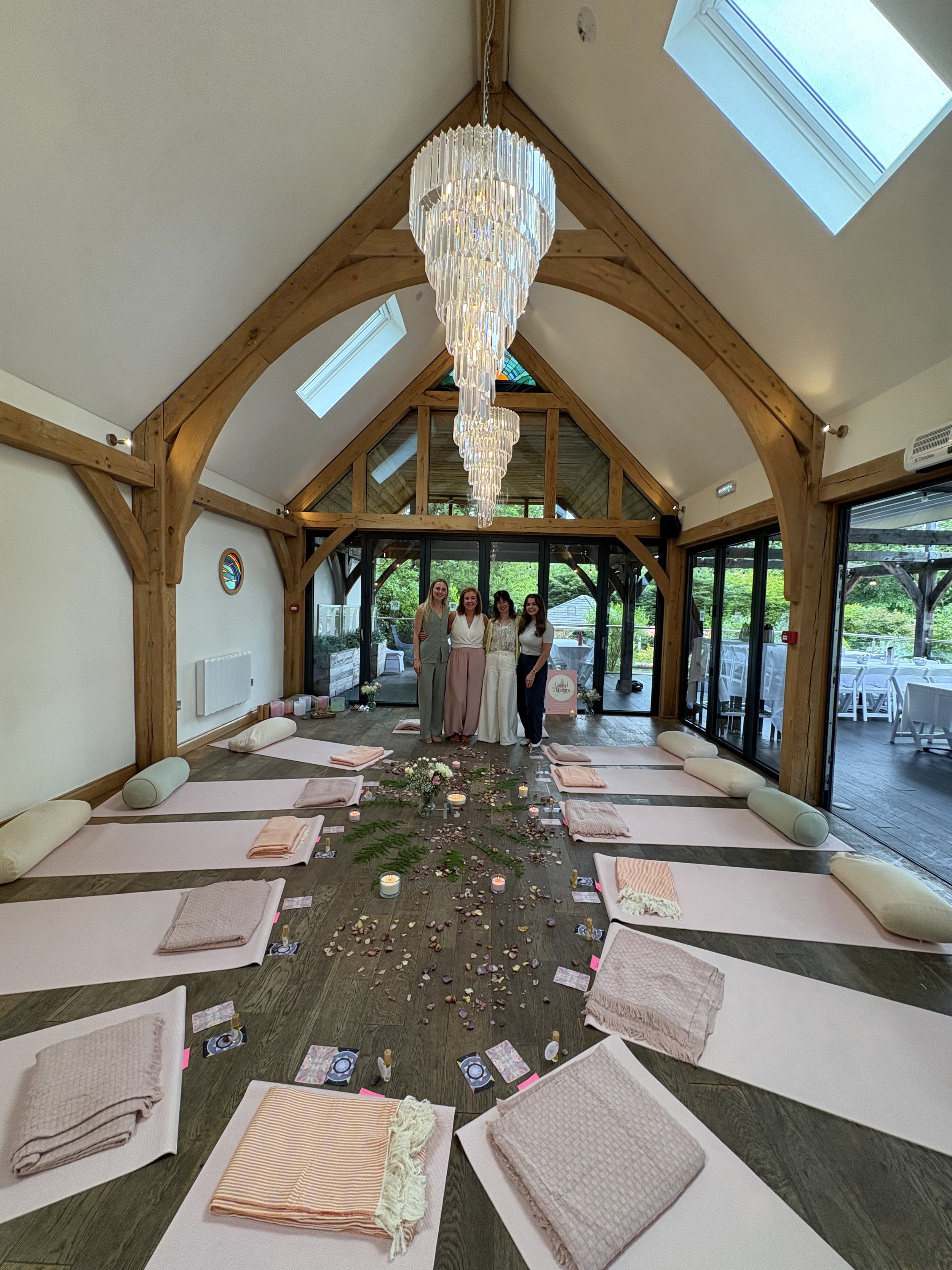 A spacious room decorated for a wedding or special event with white mats, pink towels, candles, and flowers. Four women stand in front of large windows and a door, smiling. A large, elegant chandelier hangs from the ceiling, and skylights provide natural light. The room has wooden beams and a scenic outdoor view.