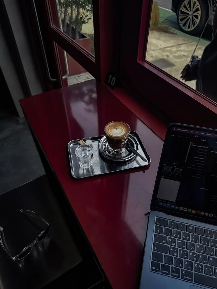 A latte with latte art in a glass cup on a black saucer on a metal tray, a small glass of water with a sugar packet, a laptop, and a pair of glasses on a black table near a window.