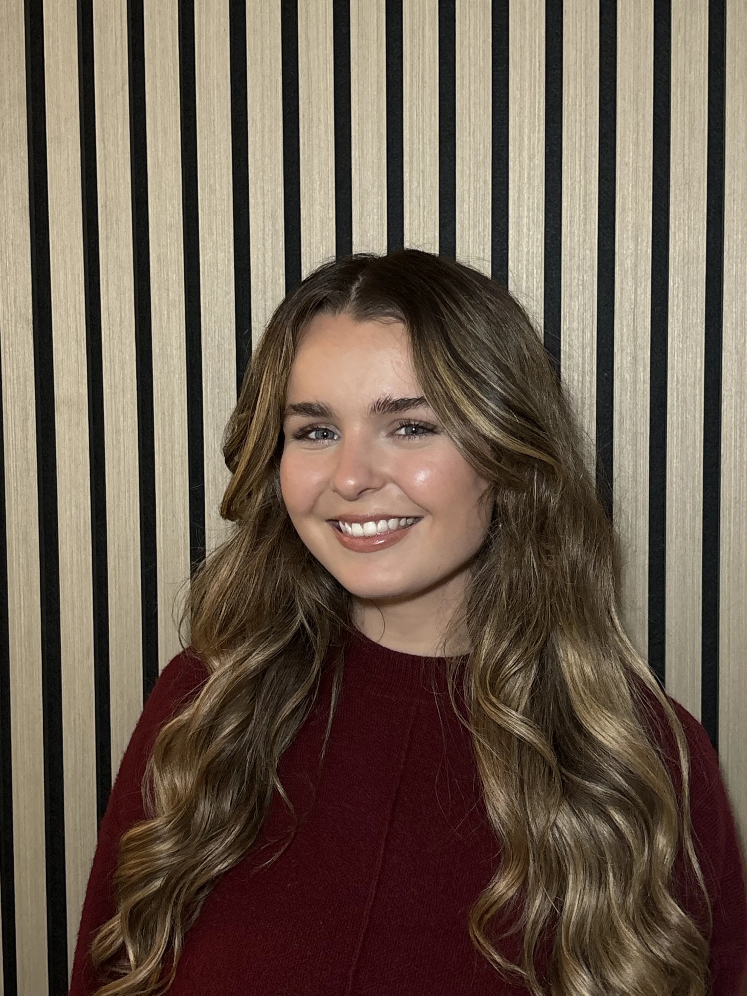 A young woman with long, wavy brown hair and blue eyes, smiling, wearing a maroon sweater, standing in front of a striped wooden wall.