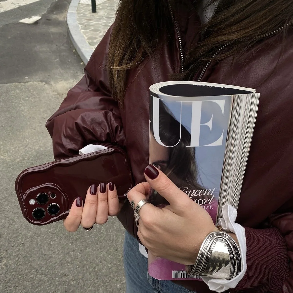 Person holding a Vogue magazine, a maroon phone case, and a smartphone, standing outdoors on a pavement with a curved stone border.