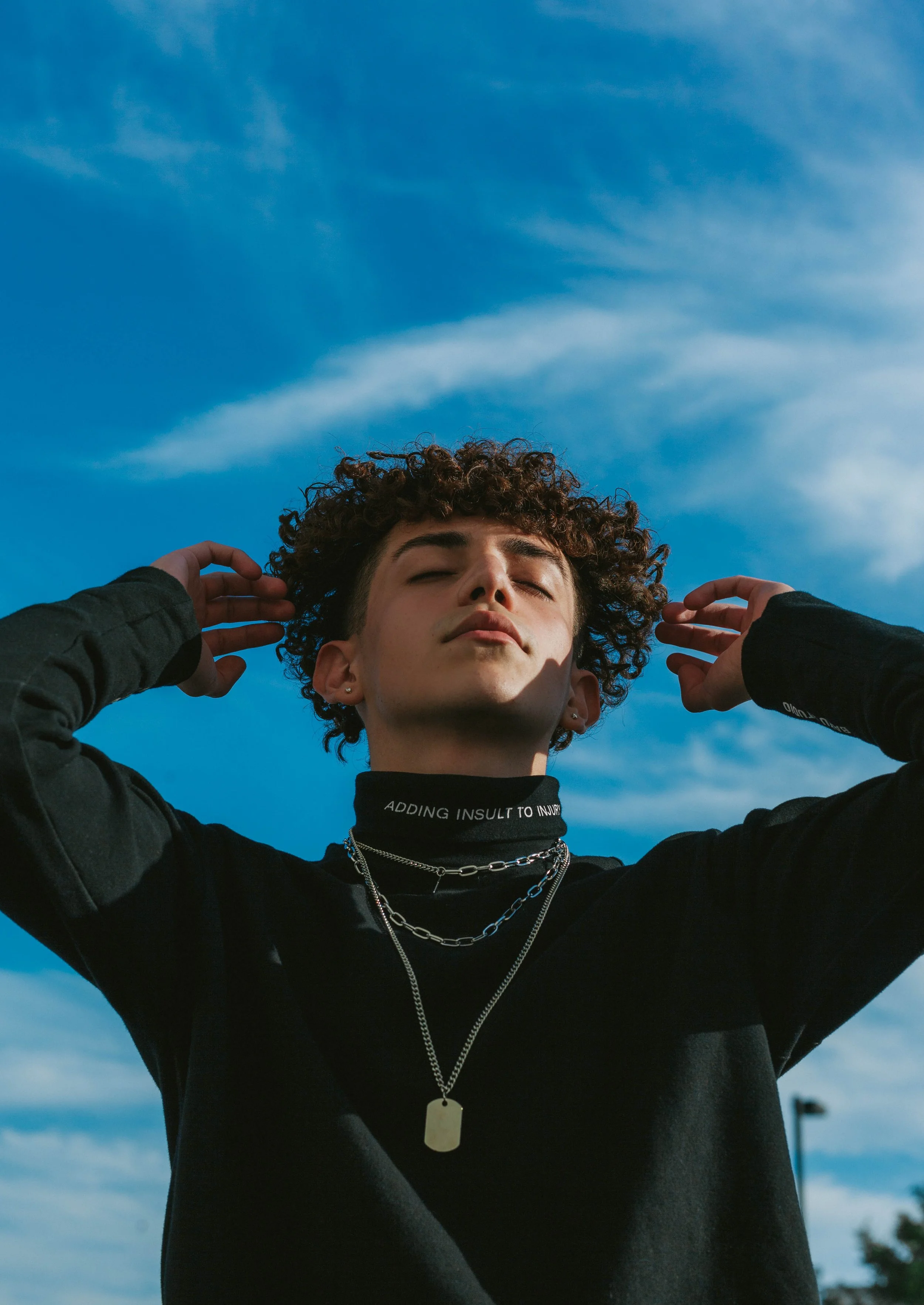 Person with short curly hair, multiple chains around neck, wearing a black top and a black neck gaiter with white text, posing outdoors against a bright blue sky with clouds.