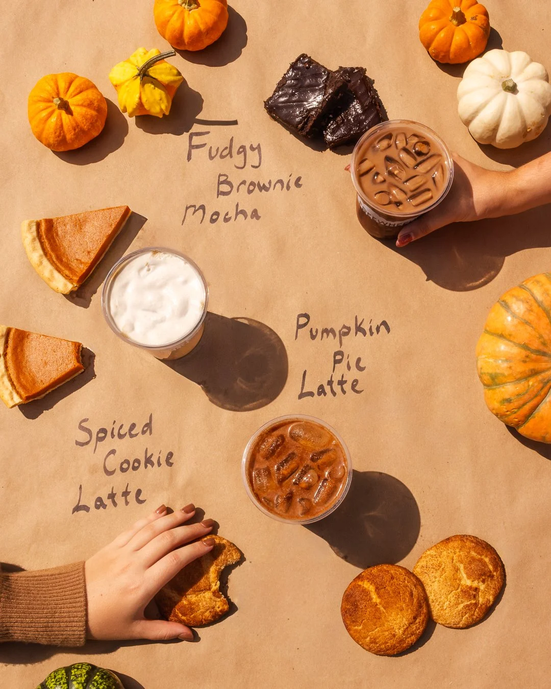Fall-themed flat lay with mini pumpkins, pumpkin pie slices, spiced cookie latte, pumpkin pie latte, and iced coffee. Hand reaching for a cookie with handwritten labels on brown paper.
