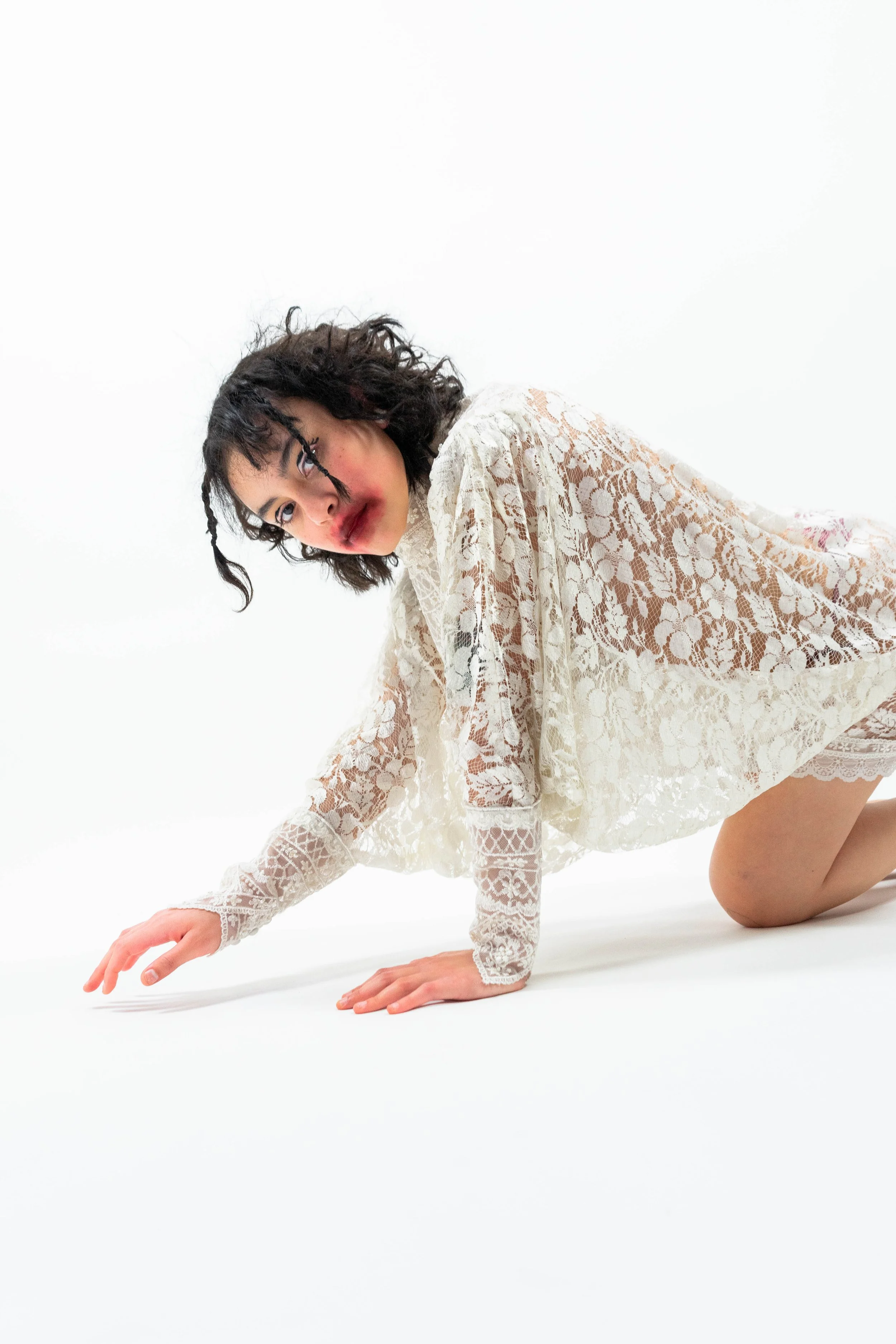 Woman in a white lace dress on hands and knees against a white background, looking at the camera.