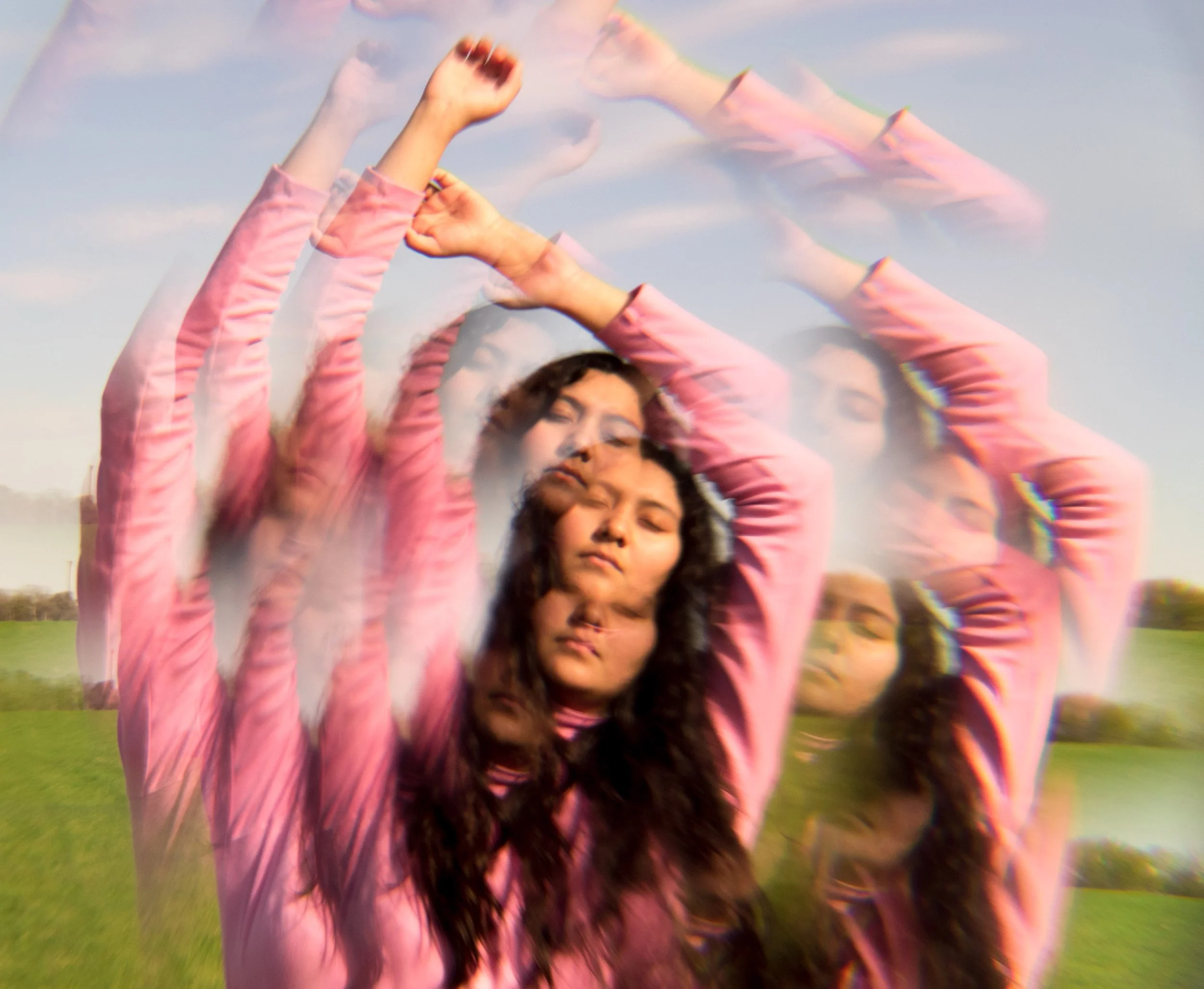 Multiple women in pink jackets with long hair raising their arms in a field with a cloudy sky.
