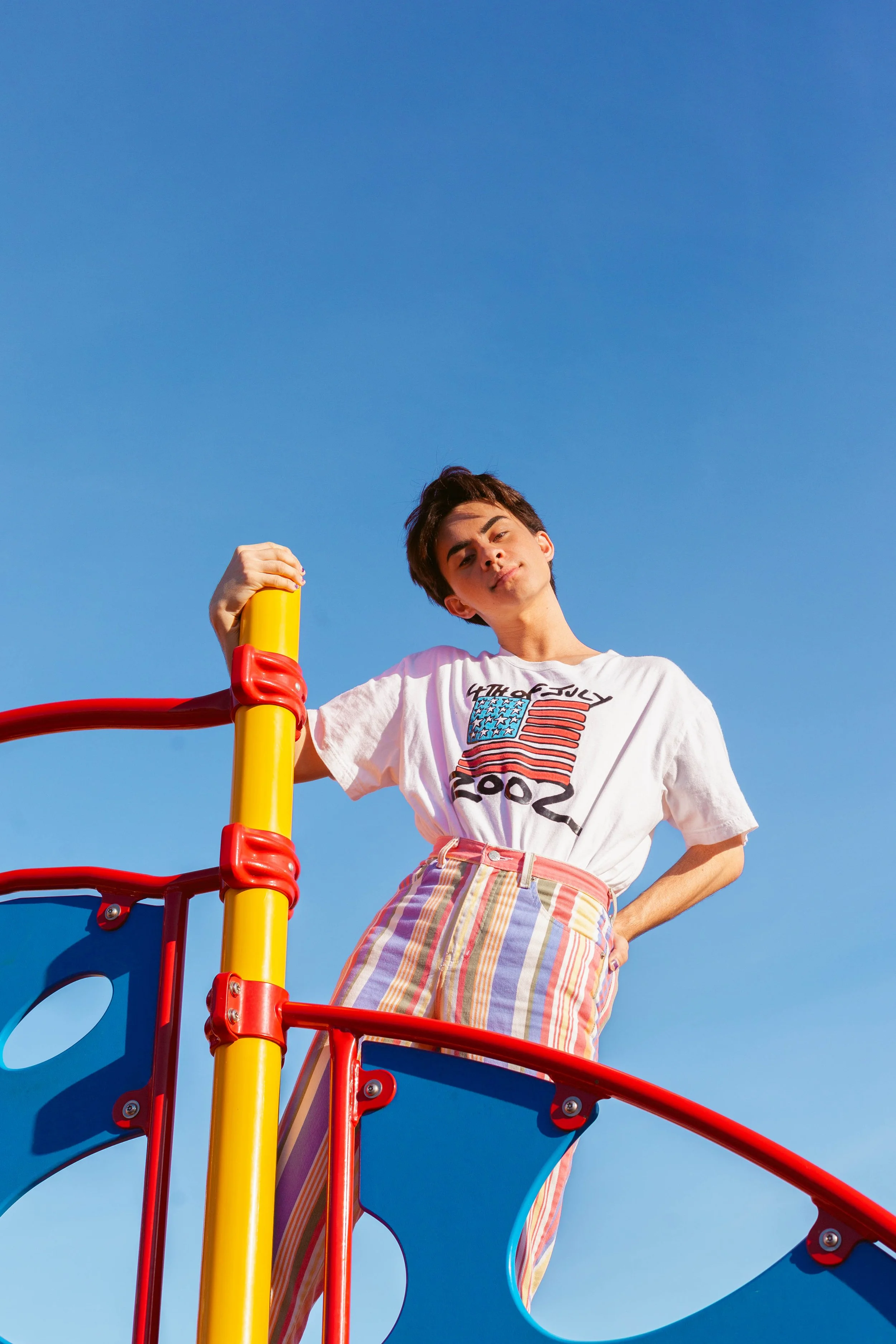 Young man on a colorful playground structure, wearing a white T-shirt with a patriotic graphic and striped pants, standing against a clear blue sky.